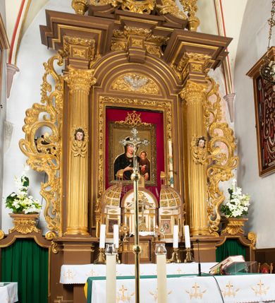 Zdjęcie nr 9: Ołtarz drewniany, architektoniczny, jednoosiowy, jednokondygnacyjny, ze zwieńczeniem, z dwiema bramkami. Jest umieszczony na murowanym, dwustopniowym podwyższeniu. Podstawa ołtarza murowana, skrzyniowa o kształcie prostopadłościanu, o obudowie drewnianej. Brak kamiennej mensy ołtarzowej. W partii antepedium nisza w kształcie prostokąta leżącego. Na mensie ustawione tabernakulum w formie centralnego kościółka zwieńczonego kopułą z latarnią.

Retabulum jest flankowane dwiema złoconymi kolumnami kompozytowymi, ustawionymi na profilowanych cokołach. Trzony kolumn są kanelowane, ozdobione w połowie wysokości główkami aniołków ujętymi dekoracją chrząstkową. Kolumny dźwigają belkowanie z wydatnym, przełamującym się nad nimi gzymsem, ozdobione ornamentem małżowinowym. W polu środkowym ołtarza profilowana, złocona i malowana rama z górną krawędzią w formie arkady z kluczem, dolna krawędź zaakcentowana uskokiem. W niej druga rama, prostokątna, ozdobiona ornamentem małżowinowym i złocona, z obrazem przedstawiającym Św. Annę Samotrzeć w otoczeniu świętych. We wnęce, za zasuwą, widnieje obraz Matki Boskiej z Dzieciątkiem w prostokątnej ramie, rzeźbionej i złoconej, pokrytej ornamentem roślinnym. Wnęka pokryta ciemnoczerwonym aksamitem, poniżej obrazu zawieszone wota. W polu arkady, w półkolistym tympanonie, na tle złoconych obłoków srebrna gołębica Ducha Świętego. Na gzymsie, na osi kolumn wazony z kwiatami. Po bokach retabulum bogato rzeźbione i złocone uszy z bujnym ornamentem małżowinowym. W zwieńczeniu, w owalnej, profilowanej i złoconej ramie obraz przedstawiający Wniebowzięcie Najświętszej Marii Panny. Rama otoczona szeroko rozbudowaną ornamentyką małżowinową. Po bokach ołtarza bramki prowadzące za ołtarz, o kształcie prostokąta stojącego, ze zwieńczeniem w formie łuku kotarowego zamkniętego od góry prostym odcinkiem, z profilowanym cokołem (pod figurę) z podwieszonym ornamentem w kształcie muszli. Górne narożniki bramek podkreślone złoconym ornamentem w kształcie wolut z elementem roślinnym. Otwór wejściowy, w kształcie analogicznym do bramki, z profilowanymi krawędziami, zakryty kotarą z aksamitu w kolorze ciemnoczerwonym. Struktura ołtarza polichromowana w kolorze ciemnobrązowym. Kolumny, pilastry, profilowania i dekoracja rzeźbiarska złocone.