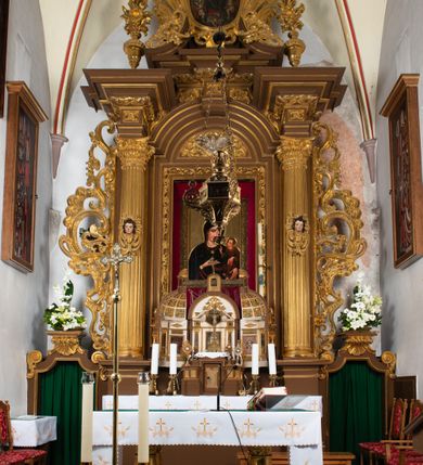 Zdjęcie nr 7: Ołtarz drewniany, architektoniczny, jednoosiowy, jednokondygnacyjny, ze zwieńczeniem, z dwiema bramkami. Jest umieszczony na murowanym, dwustopniowym podwyższeniu. Podstawa ołtarza murowana, skrzyniowa o kształcie prostopadłościanu, o obudowie drewnianej. Brak kamiennej mensy ołtarzowej. W partii antepedium nisza w kształcie prostokąta leżącego. Na mensie ustawione tabernakulum w formie centralnego kościółka zwieńczonego kopułą z latarnią.

Retabulum jest flankowane dwiema złoconymi kolumnami kompozytowymi, ustawionymi na profilowanych cokołach. Trzony kolumn są kanelowane, ozdobione w połowie wysokości główkami aniołków ujętymi dekoracją chrząstkową. Kolumny dźwigają belkowanie z wydatnym, przełamującym się nad nimi gzymsem, ozdobione ornamentem małżowinowym. W polu środkowym ołtarza profilowana, złocona i malowana rama z górną krawędzią w formie arkady z kluczem, dolna krawędź zaakcentowana uskokiem. W niej druga rama, prostokątna, ozdobiona ornamentem małżowinowym i złocona, z obrazem przedstawiającym Św. Annę Samotrzeć w otoczeniu świętych. We wnęce, za zasuwą, widnieje obraz Matki Boskiej z Dzieciątkiem w prostokątnej ramie, rzeźbionej i złoconej, pokrytej ornamentem roślinnym. Wnęka pokryta ciemnoczerwonym aksamitem, poniżej obrazu zawieszone wota. W polu arkady, w półkolistym tympanonie, na tle złoconych obłoków srebrna gołębica Ducha Świętego. Na gzymsie, na osi kolumn wazony z kwiatami. Po bokach retabulum bogato rzeźbione i złocone uszy z bujnym ornamentem małżowinowym. W zwieńczeniu, w owalnej, profilowanej i złoconej ramie obraz przedstawiający Wniebowzięcie Najświętszej Marii Panny. Rama otoczona szeroko rozbudowaną ornamentyką małżowinową. Po bokach ołtarza bramki prowadzące za ołtarz, o kształcie prostokąta stojącego, ze zwieńczeniem w formie łuku kotarowego zamkniętego od góry prostym odcinkiem, z profilowanym cokołem (pod figurę) z podwieszonym ornamentem w kształcie muszli. Górne narożniki bramek podkreślone złoconym ornamentem w kształcie wolut z elementem roślinnym. Otwór wejściowy, w kształcie analogicznym do bramki, z profilowanymi krawędziami, zakryty kotarą z aksamitu w kolorze ciemnoczerwonym. Struktura ołtarza polichromowana w kolorze ciemnobrązowym. Kolumny, pilastry, profilowania i dekoracja rzeźbiarska złocone.