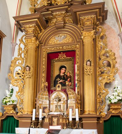 Zdjęcie nr 18: Ołtarz drewniany, architektoniczny, jednoosiowy, jednokondygnacyjny, ze zwieńczeniem, z dwiema bramkami. Jest umieszczony na murowanym, dwustopniowym podwyższeniu. Podstawa ołtarza murowana, skrzyniowa o kształcie prostopadłościanu, o obudowie drewnianej. Brak kamiennej mensy ołtarzowej. W partii antepedium nisza w kształcie prostokąta leżącego. Na mensie ustawione tabernakulum w formie centralnego kościółka zwieńczonego kopułą z latarnią.

Retabulum jest flankowane dwiema złoconymi kolumnami kompozytowymi, ustawionymi na profilowanych cokołach. Trzony kolumn są kanelowane, ozdobione w połowie wysokości główkami aniołków ujętymi dekoracją chrząstkową. Kolumny dźwigają belkowanie z wydatnym, przełamującym się nad nimi gzymsem, ozdobione ornamentem małżowinowym. W polu środkowym ołtarza profilowana, złocona i malowana rama z górną krawędzią w formie arkady z kluczem, dolna krawędź zaakcentowana uskokiem. W niej druga rama, prostokątna, ozdobiona ornamentem małżowinowym i złocona, z obrazem przedstawiającym Św. Annę Samotrzeć w otoczeniu świętych. We wnęce, za zasuwą, widnieje obraz Matki Boskiej z Dzieciątkiem w prostokątnej ramie, rzeźbionej i złoconej, pokrytej ornamentem roślinnym. Wnęka pokryta ciemnoczerwonym aksamitem, poniżej obrazu zawieszone wota. W polu arkady, w półkolistym tympanonie, na tle złoconych obłoków srebrna gołębica Ducha Świętego. Na gzymsie, na osi kolumn wazony z kwiatami. Po bokach retabulum bogato rzeźbione i złocone uszy z bujnym ornamentem małżowinowym. W zwieńczeniu, w owalnej, profilowanej i złoconej ramie obraz przedstawiający Wniebowzięcie Najświętszej Marii Panny. Rama otoczona szeroko rozbudowaną ornamentyką małżowinową. Po bokach ołtarza bramki prowadzące za ołtarz, o kształcie prostokąta stojącego, ze zwieńczeniem w formie łuku kotarowego zamkniętego od góry prostym odcinkiem, z profilowanym cokołem (pod figurę) z podwieszonym ornamentem w kształcie muszli. Górne narożniki bramek podkreślone złoconym ornamentem w kształcie wolut z elementem roślinnym. Otwór wejściowy, w kształcie analogicznym do bramki, z profilowanymi krawędziami, zakryty kotarą z aksamitu w kolorze ciemnoczerwonym. Struktura ołtarza polichromowana w kolorze ciemnobrązowym. Kolumny, pilastry, profilowania i dekoracja rzeźbiarska złocone.