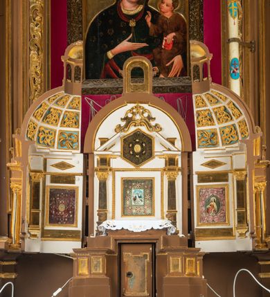 Zdjęcie nr 36: Ołtarz drewniany, architektoniczny, jednoosiowy, jednokondygnacyjny, ze zwieńczeniem, z dwiema bramkami. Jest umieszczony na murowanym, dwustopniowym podwyższeniu. Podstawa ołtarza murowana, skrzyniowa o kształcie prostopadłościanu, o obudowie drewnianej. Brak kamiennej mensy ołtarzowej. W partii antepedium nisza w kształcie prostokąta leżącego. Na mensie ustawione tabernakulum w formie centralnego kościółka zwieńczonego kopułą z latarnią.

Retabulum jest flankowane dwiema złoconymi kolumnami kompozytowymi, ustawionymi na profilowanych cokołach. Trzony kolumn są kanelowane, ozdobione w połowie wysokości główkami aniołków ujętymi dekoracją chrząstkową. Kolumny dźwigają belkowanie z wydatnym, przełamującym się nad nimi gzymsem, ozdobione ornamentem małżowinowym. W polu środkowym ołtarza profilowana, złocona i malowana rama z górną krawędzią w formie arkady z kluczem, dolna krawędź zaakcentowana uskokiem. W niej druga rama, prostokątna, ozdobiona ornamentem małżowinowym i złocona, z obrazem przedstawiającym Św. Annę Samotrzeć w otoczeniu świętych. We wnęce, za zasuwą, widnieje obraz Matki Boskiej z Dzieciątkiem w prostokątnej ramie, rzeźbionej i złoconej, pokrytej ornamentem roślinnym. Wnęka pokryta ciemnoczerwonym aksamitem, poniżej obrazu zawieszone wota. W polu arkady, w półkolistym tympanonie, na tle złoconych obłoków srebrna gołębica Ducha Świętego. Na gzymsie, na osi kolumn wazony z kwiatami. Po bokach retabulum bogato rzeźbione i złocone uszy z bujnym ornamentem małżowinowym. W zwieńczeniu, w owalnej, profilowanej i złoconej ramie obraz przedstawiający Wniebowzięcie Najświętszej Marii Panny. Rama otoczona szeroko rozbudowaną ornamentyką małżowinową. Po bokach ołtarza bramki prowadzące za ołtarz, o kształcie prostokąta stojącego, ze zwieńczeniem w formie łuku kotarowego zamkniętego od góry prostym odcinkiem, z profilowanym cokołem (pod figurę) z podwieszonym ornamentem w kształcie muszli. Górne narożniki bramek podkreślone złoconym ornamentem w kształcie wolut z elementem roślinnym. Otwór wejściowy, w kształcie analogicznym do bramki, z profilowanymi krawędziami, zakryty kotarą z aksamitu w kolorze ciemnoczerwonym. Struktura ołtarza polichromowana w kolorze ciemnobrązowym. Kolumny, pilastry, profilowania i dekoracja rzeźbiarska złocone.