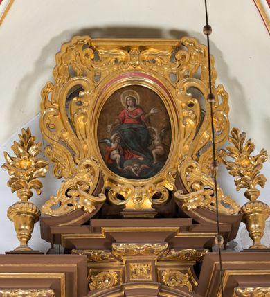 Zdjęcie nr 4: Obraz umieszczony w zwieńczeniu ołtarza głównego, w owalnej profilowanej, złoconej i malowanej ramie, ujętej ornamentem małżowinowym. W polu obrazu o kształcie owalu scena Wniebowzięcia Najświętszej Marii Panny. Dominantę kompozycji wpisanej w trójkąt stanowi ukazana frontalnie postać Marii unoszona ku niebu przez dwa nagie, antytetycznie przedstawione anioły. Pod stopami Marii i aniołów kłębią się ciemne obłoki, na ich tle, na osi kompozycji, widnieje przedstawienie cherubina. Głowa Marii nieznacznie uniesiona. Jej twarz jest okrągła, oczy duże, wzrok wzniesiony ku górze, wyraz twarzy pogodny, z delikatnym uśmiechem. Na głowie Maria ma chustę w kolorze jasnej ochry, spływającą wąską fałdą wokół szyi i przez pierś na lewe ramię, zaś wokół niej nimb składający się z promienistej glorii i złotego otoku. Ręce Marii są rozłożone, dłonie ukazane wewnętrzną stroną. Prawa ręka wyprostowana, skierowana w dół, lewa, zgięta w łokciu, skierowana ku górze. Spod sukni Marii wystaje prawa stopa z zaznaczonym konturem sandała. Madonna jest ubrana w wysoko przepasaną czerwoną suknię, z podwiniętymi szerokimi rękawami, spod których wystają wąskie długie rękawy spodniej szaty w kolorze jasnej ochry oraz ciemnoniebieski, szeroki płaszcz okrywający jej kolana. Fałdy płaszcza zaznaczone  łamiącymi się liniami.  
Obejmujące kolana Marii i zwrócone ku niej anioły są nagie, przepasane jedynie szarfami: anioł z ciemnoniebieskim skrzydłem, po prawej stronie Matki Boskiej – szarfą czerwoną, przełożoną przez lewe ramię do prawego boku i związaną wydatnym węzłem, anioł z ciemnozielonym skrzydłem, po lewej stronie Marii – szarfą w kolorze jasnej ochry, przewiązaną w pasie, w węzłem na plecach. 
Tło monochromatyczne, w odcieniach jasnego brązu i ochry. Kontury postaci podkreślone ciemnym brązem i czernią. Karnacje twarzy naturalne, modelowane światłocieniowo, policzki podkreślone różem. Włosy Marii i aniołów w kolorze jasnego brązu. Skrzydła aniołów i cherubina modelowane bielą. Kolorystyka obrazu stonowana, ciepła, nasycona. Dominantę kolorystyczną stanowi czerwień i ciemny błękit szat Najświętszej Marii Panny.