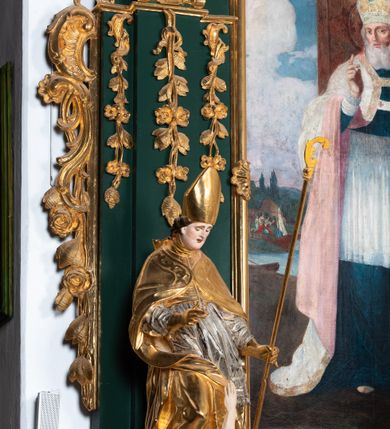 Zdjęcie nr 16: Ołtarz przyścienny, architektoniczny, jednokondygnacyjny, jednoosiowy ze zwieńczeniem. Umieszczony na dwustopniowym, kamiennym podwyższeniu. Mensa prostopadłościenna, z portatylem i współczesnym antepedium ozdobionym postacią św. Weroniki trzymającej chustę z odbiciem twarzy Chrystusa (veraikon) i rzeźbionym ornamentem ze srebrzonych liści i złoconych kiści winogron; tło w kolorze struktury ołtarza. Retabulum na wysokim profilowanym, dwustopniowym cokole, flankowane zwielokrotnionymi pilastrami z jońskimi kapitelami. Pilastry dźwigają przerwane belkowanie z wydatnym profilowanym gzymsem z przerwanym przyczółkiem. Zwieńczenie o wklęsłych bokach ujęte spływami wolutowymi dźwigającymi gzyms w formie nadwieszonego łuku odcinkowego. Na tle pilastrów ustawione na profilowanych impostach rzeźby: po prawej stronie św. Wojciecha, po lewej św. Stanisława. Na przyczółkach znajdują się siedzące putta. Pole środkowe o kształcie prostokąta stojącego od góry zamkniętego łukiem półkolistym nadwieszonym, w kluczu ornament z motywu rocaille. Wewnątrz pola ujęty profilowaną, złoconą ramą obraz z przedstawieniem św. Mikołaja z Miry. W zwieńczeniu obraz w ramie z wklęsłymi bokami i wypukłymi dolną i górną krawędzią przedstawiający św. Marię Magdalenę. Nastawę ujmują ornamentalne uszy, utworzone z motywów rocaille'owych i liściasto-kwiatowych zwisów. Na kapitele są nałożone rocaille'owe kartusze, a pod nimi podwieszone zwisające ulistnione i ukwiecone gałązki wzbogacone motywami rocaille'ów. Struktura ołtarza polichromowana w kolorze ciemnozielonym. Bazy pilastrów, kapitele, uszy, ornamenty, profilowania, obramienia płycin i ramy obrazów złocone. Rzeźby świętych złocone, srebrzone i polichromowane. Rzeźby aniołków złocone i polichromowane.

