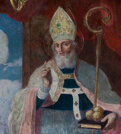 Zdjęcie nr 1: Obraz w kształcie stojącego prostokąta zamkniętego od góry półkolistym łukiem, nadwieszonym, w profilowanej, złoconej ramie ozdobionej motywem muszli w połowie długości dłuższych boków. Został umieszczony w polu środkowym ołtarza bocznego prawego. Postać św. Mikołaja ukazana w pozycji stojącej, frontalnie, we wnętrzu na tle upiętej ciemnoczerwonej draperii. Głowa świętego przechylona nieznacznie w prawo. Twarz owalna, okolona długą, siwą brodą. Oczy duże, głęboko osadzone, wzrok skierowany na wprost. Nos wydatny, prosty, usta zaczerwienione. Na głowie złota infuła, ozdobiona kwadratowym zielonym kamieniem szlachetnym. Wokół głowy nimb. Święty unosi prawą rękę w geście błogosławieństwa, w lewej ręce, o którą oparty jest pastorał, trzyma księgę, z leżącymi na niej trzema złotymi kulami. Ubrany jest w granatową sutannę i białą komżę, układającą się w drobne płytkie fałdy. Na piersi ma paliusz, a na ramionach kapę w kolorze szarym o różowej podszewce. Z lewej strony obrazu okno, w nim widoczny pejzaż z rzeką. Nad rzeką most, przy nim św. Mikołaj wręczający trzem pannom sakiewki z posagiem. Na brzegu rzeki łódź, w głębi widoczny zarys architektury, drzewa i zarysy wzgórz, ponad którymi na błękitnym niebie kłębią się białe chmury. Po prawej stronie obrazu stolik nakryty ciemnobłękitną tkaniną, na nim taca z kwiatami, różańcem i figurką. Kolorystyka obrazu stonowana z silnym akcentem bieli komży i różu podszewki płaszcza.
