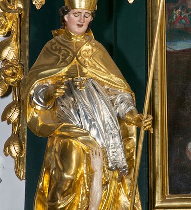 Zdjęcie nr 15: Pełnoplastyczna rzeźba drewniana przedstawiająca św. Stanisława, przyścienna, polichromowana, złocona i srebrzona, wydrążona z tyłu. Ustawiona na niskiej prostokątnej podstawie. Święty ukazany całopostaciowo, w pozycji stojącej w kontrapoście, z prawą nogą ugiętą w kolanie i odstawioną w bok, z wyraźnym przegięciem ciała w lewo i skręceniem korpusu w prawo. W lewej ręce pastorał trzymany na wysokości bioder, prawa zgięta w łokciu i uniesiona przed siebie. Głowa mała, osadzona na długiej szyi, pochylona w dół, lekko zwrócona w lewo. Twarz owalna, policzki pełne. Oczy prawie całkowicie przesłonięte powiekami, wzrok skierowany w dół. Nos duży, prosty, usta małe, pełne, o uniesionych w górę kącikach. Głowę po bokach okalają krótkie włosy układające się w wywinięte na zewnątrz pukle. Na głowie infuła ozdobiona krzyżem równoramiennym o lekko rozszerzających się ramionach, ze wstęgami na plecach. Święty ubrany w albę, rokietę z szeroką, dekoracyjną koronką i mucet. Na ramiona narzucona kapa z galonem ozdobionym stylizowanym motywem roślinnym. Na piersi widoczny pektorał, na szyi humerał, na dłoniach rękawiczki. Spod szat widoczny lewy but. Szaty świętego ukształtowane ekspresyjnie, o ostro ciętych, skośnych fałdach. Poły płaszcza wywinięte na zewnątrz. Odwinięta poła płaszcza pod prawą ręką tworzy obszerny fałd o kształcie litery "S". Z przodu figury, u stóp świętego postać wyłaniającego się z grobu Piotrowina, z mocno odchyloną do tyłu głową nakrytą całunem i wyprostowaną, wyciągniętą pionowo w górę ręką, z widocznie zaznaczonymi mięśniami i dłonią o długich, szczupłych palcach. Twarz Piotrowina wychudzona, z dużym, lekko garbatym nosem, ponad ustami proste wąsy.  Cały strój świętego, z wyjątkiem srebrzonej rokiety, złocony. Twarz i włosy polichromowane. Postać Piotrowina, z wyjątkiem srebrzonego całunu, polichromowana.