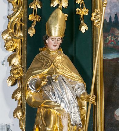 Zdjęcie nr 1: Pełnoplastyczna rzeźba drewniana przedstawiająca św. Stanisława, przyścienna, polichromowana, złocona i srebrzona, wydrążona z tyłu. Ustawiona na niskiej prostokątnej podstawie. Święty ukazany całopostaciowo, w pozycji stojącej w kontrapoście, z prawą nogą ugiętą w kolanie i odstawioną w bok, z wyraźnym przegięciem ciała w lewo i skręceniem korpusu w prawo. W lewej ręce pastorał trzymany na wysokości bioder, prawa zgięta w łokciu i uniesiona przed siebie. Głowa mała, osadzona na długiej szyi, pochylona w dół, lekko zwrócona w lewo. Twarz owalna, policzki pełne. Oczy prawie całkowicie przesłonięte powiekami, wzrok skierowany w dół. Nos duży, prosty, usta małe, pełne, o uniesionych w górę kącikach. Głowę po bokach okalają krótkie włosy układające się w wywinięte na zewnątrz pukle. Na głowie infuła ozdobiona krzyżem równoramiennym o lekko rozszerzających się ramionach, ze wstęgami na plecach. Święty ubrany w albę, rokietę z szeroką, dekoracyjną koronką i mucet. Na ramiona narzucona kapa z galonem ozdobionym stylizowanym motywem roślinnym. Na piersi widoczny pektorał, na szyi humerał, na dłoniach rękawiczki. Spod szat widoczny lewy but. Szaty świętego ukształtowane ekspresyjnie, o ostro ciętych, skośnych fałdach. Poły płaszcza wywinięte na zewnątrz. Odwinięta poła płaszcza pod prawą ręką tworzy obszerny fałd o kształcie litery "S". Z przodu figury, u stóp świętego postać wyłaniającego się z grobu Piotrowina, z mocno odchyloną do tyłu głową nakrytą całunem i wyprostowaną, wyciągniętą pionowo w górę ręką, z widocznie zaznaczonymi mięśniami i dłonią o długich, szczupłych palcach. Twarz Piotrowina wychudzona, z dużym, lekko garbatym nosem, ponad ustami proste wąsy.  Cały strój świętego, z wyjątkiem srebrzonej rokiety, złocony. Twarz i włosy polichromowane. Postać Piotrowina, z wyjątkiem srebrzonego całunu, polichromowana.