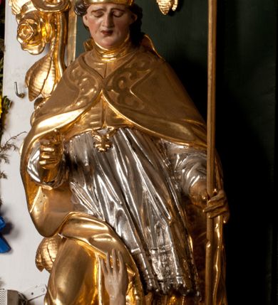 Zdjęcie nr 16: Pełnoplastyczna rzeźba drewniana przedstawiająca św. Stanisława, przyścienna, polichromowana, złocona i srebrzona, wydrążona z tyłu. Ustawiona na niskiej prostokątnej podstawie. Święty ukazany całopostaciowo, w pozycji stojącej w kontrapoście, z prawą nogą ugiętą w kolanie i odstawioną w bok, z wyraźnym przegięciem ciała w lewo i skręceniem korpusu w prawo. W lewej ręce pastorał trzymany na wysokości bioder, prawa zgięta w łokciu i uniesiona przed siebie. Głowa mała, osadzona na długiej szyi, pochylona w dół, lekko zwrócona w lewo. Twarz owalna, policzki pełne. Oczy prawie całkowicie przesłonięte powiekami, wzrok skierowany w dół. Nos duży, prosty, usta małe, pełne, o uniesionych w górę kącikach. Głowę po bokach okalają krótkie włosy układające się w wywinięte na zewnątrz pukle. Na głowie infuła ozdobiona krzyżem równoramiennym o lekko rozszerzających się ramionach, ze wstęgami na plecach. Święty ubrany w albę, rokietę z szeroką, dekoracyjną koronką i mucet. Na ramiona narzucona kapa z galonem ozdobionym stylizowanym motywem roślinnym. Na piersi widoczny pektorał, na szyi humerał, na dłoniach rękawiczki. Spod szat widoczny lewy but. Szaty świętego ukształtowane ekspresyjnie, o ostro ciętych, skośnych fałdach. Poły płaszcza wywinięte na zewnątrz. Odwinięta poła płaszcza pod prawą ręką tworzy obszerny fałd o kształcie litery &quot;S&quot;. Z przodu figury, u stóp świętego postać wyłaniającego się z grobu Piotrowina, z mocno odchyloną do tyłu głową nakrytą całunem i wyprostowaną, wyciągniętą pionowo w górę ręką, z widocznie zaznaczonymi mięśniami i dłonią o długich, szczupłych palcach. Twarz Piotrowina wychudzona, z dużym, lekko garbatym nosem, ponad ustami proste wąsy.  Cały strój świętego, z wyjątkiem srebrzonej rokiety, złocony. Twarz i włosy polichromowane. Postać Piotrowina, z wyjątkiem srebrzonego całunu, polichromowana.