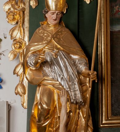 Zdjęcie nr 17: Pełnoplastyczna rzeźba drewniana przedstawiająca św. Stanisława, przyścienna, polichromowana, złocona i srebrzona, wydrążona z tyłu. Ustawiona na niskiej prostokątnej podstawie. Święty ukazany całopostaciowo, w pozycji stojącej w kontrapoście, z prawą nogą ugiętą w kolanie i odstawioną w bok, z wyraźnym przegięciem ciała w lewo i skręceniem korpusu w prawo. W lewej ręce pastorał trzymany na wysokości bioder, prawa zgięta w łokciu i uniesiona przed siebie. Głowa mała, osadzona na długiej szyi, pochylona w dół, lekko zwrócona w lewo. Twarz owalna, policzki pełne. Oczy prawie całkowicie przesłonięte powiekami, wzrok skierowany w dół. Nos duży, prosty, usta małe, pełne, o uniesionych w górę kącikach. Głowę po bokach okalają krótkie włosy układające się w wywinięte na zewnątrz pukle. Na głowie infuła ozdobiona krzyżem równoramiennym o lekko rozszerzających się ramionach, ze wstęgami na plecach. Święty ubrany w albę, rokietę z szeroką, dekoracyjną koronką i mucet. Na ramiona narzucona kapa z galonem ozdobionym stylizowanym motywem roślinnym. Na piersi widoczny pektorał, na szyi humerał, na dłoniach rękawiczki. Spod szat widoczny lewy but. Szaty świętego ukształtowane ekspresyjnie, o ostro ciętych, skośnych fałdach. Poły płaszcza wywinięte na zewnątrz. Odwinięta poła płaszcza pod prawą ręką tworzy obszerny fałd o kształcie litery "S". Z przodu figury, u stóp świętego postać wyłaniającego się z grobu Piotrowina, z mocno odchyloną do tyłu głową nakrytą całunem i wyprostowaną, wyciągniętą pionowo w górę ręką, z widocznie zaznaczonymi mięśniami i dłonią o długich, szczupłych palcach. Twarz Piotrowina wychudzona, z dużym, lekko garbatym nosem, ponad ustami proste wąsy.  Cały strój świętego, z wyjątkiem srebrzonej rokiety, złocony. Twarz i włosy polichromowane. Postać Piotrowina, z wyjątkiem srebrzonego całunu, polichromowana.