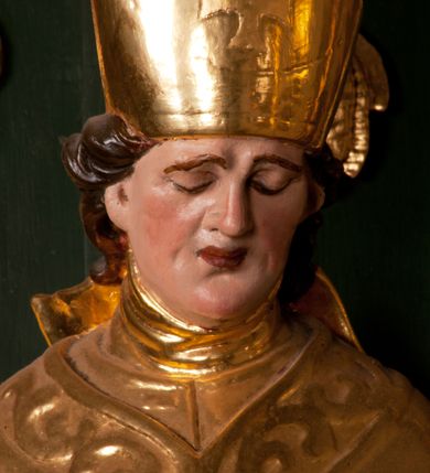Zdjęcie nr 18: Pełnoplastyczna rzeźba drewniana przedstawiająca św. Stanisława, przyścienna, polichromowana, złocona i srebrzona, wydrążona z tyłu. Ustawiona na niskiej prostokątnej podstawie. Święty ukazany całopostaciowo, w pozycji stojącej w kontrapoście, z prawą nogą ugiętą w kolanie i odstawioną w bok, z wyraźnym przegięciem ciała w lewo i skręceniem korpusu w prawo. W lewej ręce pastorał trzymany na wysokości bioder, prawa zgięta w łokciu i uniesiona przed siebie. Głowa mała, osadzona na długiej szyi, pochylona w dół, lekko zwrócona w lewo. Twarz owalna, policzki pełne. Oczy prawie całkowicie przesłonięte powiekami, wzrok skierowany w dół. Nos duży, prosty, usta małe, pełne, o uniesionych w górę kącikach. Głowę po bokach okalają krótkie włosy układające się w wywinięte na zewnątrz pukle. Na głowie infuła ozdobiona krzyżem równoramiennym o lekko rozszerzających się ramionach, ze wstęgami na plecach. Święty ubrany w albę, rokietę z szeroką, dekoracyjną koronką i mucet. Na ramiona narzucona kapa z galonem ozdobionym stylizowanym motywem roślinnym. Na piersi widoczny pektorał, na szyi humerał, na dłoniach rękawiczki. Spod szat widoczny lewy but. Szaty świętego ukształtowane ekspresyjnie, o ostro ciętych, skośnych fałdach. Poły płaszcza wywinięte na zewnątrz. Odwinięta poła płaszcza pod prawą ręką tworzy obszerny fałd o kształcie litery "S". Z przodu figury, u stóp świętego postać wyłaniającego się z grobu Piotrowina, z mocno odchyloną do tyłu głową nakrytą całunem i wyprostowaną, wyciągniętą pionowo w górę ręką, z widocznie zaznaczonymi mięśniami i dłonią o długich, szczupłych palcach. Twarz Piotrowina wychudzona, z dużym, lekko garbatym nosem, ponad ustami proste wąsy.  Cały strój świętego, z wyjątkiem srebrzonej rokiety, złocony. Twarz i włosy polichromowane. Postać Piotrowina, z wyjątkiem srebrzonego całunu, polichromowana.