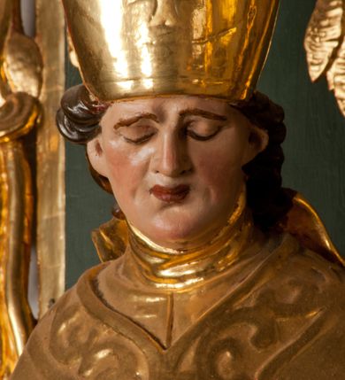 Zdjęcie nr 20: Pełnoplastyczna rzeźba drewniana przedstawiająca św. Stanisława, przyścienna, polichromowana, złocona i srebrzona, wydrążona z tyłu. Ustawiona na niskiej prostokątnej podstawie. Święty ukazany całopostaciowo, w pozycji stojącej w kontrapoście, z prawą nogą ugiętą w kolanie i odstawioną w bok, z wyraźnym przegięciem ciała w lewo i skręceniem korpusu w prawo. W lewej ręce pastorał trzymany na wysokości bioder, prawa zgięta w łokciu i uniesiona przed siebie. Głowa mała, osadzona na długiej szyi, pochylona w dół, lekko zwrócona w lewo. Twarz owalna, policzki pełne. Oczy prawie całkowicie przesłonięte powiekami, wzrok skierowany w dół. Nos duży, prosty, usta małe, pełne, o uniesionych w górę kącikach. Głowę po bokach okalają krótkie włosy układające się w wywinięte na zewnątrz pukle. Na głowie infuła ozdobiona krzyżem równoramiennym o lekko rozszerzających się ramionach, ze wstęgami na plecach. Święty ubrany w albę, rokietę z szeroką, dekoracyjną koronką i mucet. Na ramiona narzucona kapa z galonem ozdobionym stylizowanym motywem roślinnym. Na piersi widoczny pektorał, na szyi humerał, na dłoniach rękawiczki. Spod szat widoczny lewy but. Szaty świętego ukształtowane ekspresyjnie, o ostro ciętych, skośnych fałdach. Poły płaszcza wywinięte na zewnątrz. Odwinięta poła płaszcza pod prawą ręką tworzy obszerny fałd o kształcie litery "S". Z przodu figury, u stóp świętego postać wyłaniającego się z grobu Piotrowina, z mocno odchyloną do tyłu głową nakrytą całunem i wyprostowaną, wyciągniętą pionowo w górę ręką, z widocznie zaznaczonymi mięśniami i dłonią o długich, szczupłych palcach. Twarz Piotrowina wychudzona, z dużym, lekko garbatym nosem, ponad ustami proste wąsy.  Cały strój świętego, z wyjątkiem srebrzonej rokiety, złocony. Twarz i włosy polichromowane. Postać Piotrowina, z wyjątkiem srebrzonego całunu, polichromowana.