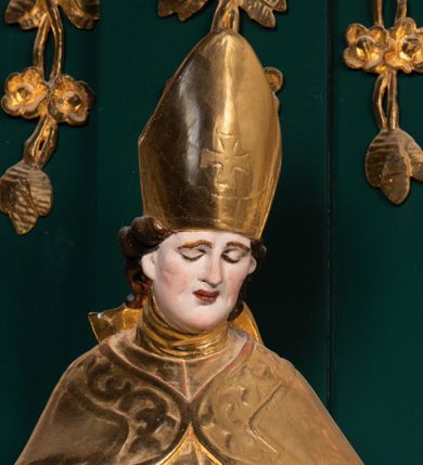 Zdjęcie nr 21: Pełnoplastyczna rzeźba drewniana przedstawiająca św. Stanisława, przyścienna, polichromowana, złocona i srebrzona, wydrążona z tyłu. Ustawiona na niskiej prostokątnej podstawie. Święty ukazany całopostaciowo, w pozycji stojącej w kontrapoście, z prawą nogą ugiętą w kolanie i odstawioną w bok, z wyraźnym przegięciem ciała w lewo i skręceniem korpusu w prawo. W lewej ręce pastorał trzymany na wysokości bioder, prawa zgięta w łokciu i uniesiona przed siebie. Głowa mała, osadzona na długiej szyi, pochylona w dół, lekko zwrócona w lewo. Twarz owalna, policzki pełne. Oczy prawie całkowicie przesłonięte powiekami, wzrok skierowany w dół. Nos duży, prosty, usta małe, pełne, o uniesionych w górę kącikach. Głowę po bokach okalają krótkie włosy układające się w wywinięte na zewnątrz pukle. Na głowie infuła ozdobiona krzyżem równoramiennym o lekko rozszerzających się ramionach, ze wstęgami na plecach. Święty ubrany w albę, rokietę z szeroką, dekoracyjną koronką i mucet. Na ramiona narzucona kapa z galonem ozdobionym stylizowanym motywem roślinnym. Na piersi widoczny pektorał, na szyi humerał, na dłoniach rękawiczki. Spod szat widoczny lewy but. Szaty świętego ukształtowane ekspresyjnie, o ostro ciętych, skośnych fałdach. Poły płaszcza wywinięte na zewnątrz. Odwinięta poła płaszcza pod prawą ręką tworzy obszerny fałd o kształcie litery &quot;S&quot;. Z przodu figury, u stóp świętego postać wyłaniającego się z grobu Piotrowina, z mocno odchyloną do tyłu głową nakrytą całunem i wyprostowaną, wyciągniętą pionowo w górę ręką, z widocznie zaznaczonymi mięśniami i dłonią o długich, szczupłych palcach. Twarz Piotrowina wychudzona, z dużym, lekko garbatym nosem, ponad ustami proste wąsy.  Cały strój świętego, z wyjątkiem srebrzonej rokiety, złocony. Twarz i włosy polichromowane. Postać Piotrowina, z wyjątkiem srebrzonego całunu, polichromowana.