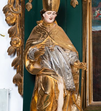 Zdjęcie nr 5: Pełnoplastyczna rzeźba drewniana przedstawiająca św. Stanisława, przyścienna, polichromowana, złocona i srebrzona, wydrążona z tyłu. Ustawiona na niskiej prostokątnej podstawie. Święty ukazany całopostaciowo, w pozycji stojącej w kontrapoście, z prawą nogą ugiętą w kolanie i odstawioną w bok, z wyraźnym przegięciem ciała w lewo i skręceniem korpusu w prawo. W lewej ręce pastorał trzymany na wysokości bioder, prawa zgięta w łokciu i uniesiona przed siebie. Głowa mała, osadzona na długiej szyi, pochylona w dół, lekko zwrócona w lewo. Twarz owalna, policzki pełne. Oczy prawie całkowicie przesłonięte powiekami, wzrok skierowany w dół. Nos duży, prosty, usta małe, pełne, o uniesionych w górę kącikach. Głowę po bokach okalają krótkie włosy układające się w wywinięte na zewnątrz pukle. Na głowie infuła ozdobiona krzyżem równoramiennym o lekko rozszerzających się ramionach, ze wstęgami na plecach. Święty ubrany w albę, rokietę z szeroką, dekoracyjną koronką i mucet. Na ramiona narzucona kapa z galonem ozdobionym stylizowanym motywem roślinnym. Na piersi widoczny pektorał, na szyi humerał, na dłoniach rękawiczki. Spod szat widoczny lewy but. Szaty świętego ukształtowane ekspresyjnie, o ostro ciętych, skośnych fałdach. Poły płaszcza wywinięte na zewnątrz. Odwinięta poła płaszcza pod prawą ręką tworzy obszerny fałd o kształcie litery "S". Z przodu figury, u stóp świętego postać wyłaniającego się z grobu Piotrowina, z mocno odchyloną do tyłu głową nakrytą całunem i wyprostowaną, wyciągniętą pionowo w górę ręką, z widocznie zaznaczonymi mięśniami i dłonią o długich, szczupłych palcach. Twarz Piotrowina wychudzona, z dużym, lekko garbatym nosem, ponad ustami proste wąsy.  Cały strój świętego, z wyjątkiem srebrzonej rokiety, złocony. Twarz i włosy polichromowane. Postać Piotrowina, z wyjątkiem srebrzonego całunu, polichromowana.