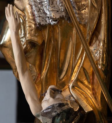 Zdjęcie nr 3: Pełnoplastyczna rzeźba drewniana przedstawiająca św. Stanisława, przyścienna, polichromowana, złocona i srebrzona, wydrążona z tyłu. Ustawiona na niskiej prostokątnej podstawie. Święty ukazany całopostaciowo, w pozycji stojącej w kontrapoście, z prawą nogą ugiętą w kolanie i odstawioną w bok, z wyraźnym przegięciem ciała w lewo i skręceniem korpusu w prawo. W lewej ręce pastorał trzymany na wysokości bioder, prawa zgięta w łokciu i uniesiona przed siebie. Głowa mała, osadzona na długiej szyi, pochylona w dół, lekko zwrócona w lewo. Twarz owalna, policzki pełne. Oczy prawie całkowicie przesłonięte powiekami, wzrok skierowany w dół. Nos duży, prosty, usta małe, pełne, o uniesionych w górę kącikach. Głowę po bokach okalają krótkie włosy układające się w wywinięte na zewnątrz pukle. Na głowie infuła ozdobiona krzyżem równoramiennym o lekko rozszerzających się ramionach, ze wstęgami na plecach. Święty ubrany w albę, rokietę z szeroką, dekoracyjną koronką i mucet. Na ramiona narzucona kapa z galonem ozdobionym stylizowanym motywem roślinnym. Na piersi widoczny pektorał, na szyi humerał, na dłoniach rękawiczki. Spod szat widoczny lewy but. Szaty świętego ukształtowane ekspresyjnie, o ostro ciętych, skośnych fałdach. Poły płaszcza wywinięte na zewnątrz. Odwinięta poła płaszcza pod prawą ręką tworzy obszerny fałd o kształcie litery "S". Z przodu figury, u stóp świętego postać wyłaniającego się z grobu Piotrowina, z mocno odchyloną do tyłu głową nakrytą całunem i wyprostowaną, wyciągniętą pionowo w górę ręką, z widocznie zaznaczonymi mięśniami i dłonią o długich, szczupłych palcach. Twarz Piotrowina wychudzona, z dużym, lekko garbatym nosem, ponad ustami proste wąsy.  Cały strój świętego, z wyjątkiem srebrzonej rokiety, złocony. Twarz i włosy polichromowane. Postać Piotrowina, z wyjątkiem srebrzonego całunu, polichromowana.