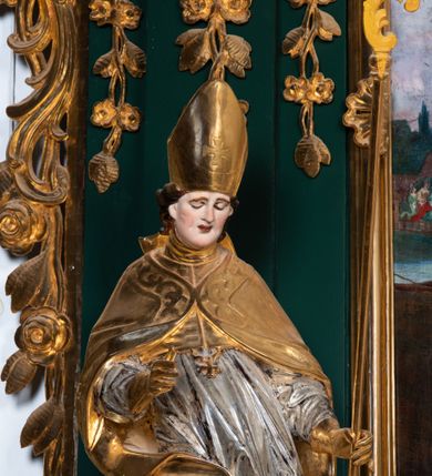 Zdjęcie nr 4: Pełnoplastyczna rzeźba drewniana przedstawiająca św. Stanisława, przyścienna, polichromowana, złocona i srebrzona, wydrążona z tyłu. Ustawiona na niskiej prostokątnej podstawie. Święty ukazany całopostaciowo, w pozycji stojącej w kontrapoście, z prawą nogą ugiętą w kolanie i odstawioną w bok, z wyraźnym przegięciem ciała w lewo i skręceniem korpusu w prawo. W lewej ręce pastorał trzymany na wysokości bioder, prawa zgięta w łokciu i uniesiona przed siebie. Głowa mała, osadzona na długiej szyi, pochylona w dół, lekko zwrócona w lewo. Twarz owalna, policzki pełne. Oczy prawie całkowicie przesłonięte powiekami, wzrok skierowany w dół. Nos duży, prosty, usta małe, pełne, o uniesionych w górę kącikach. Głowę po bokach okalają krótkie włosy układające się w wywinięte na zewnątrz pukle. Na głowie infuła ozdobiona krzyżem równoramiennym o lekko rozszerzających się ramionach, ze wstęgami na plecach. Święty ubrany w albę, rokietę z szeroką, dekoracyjną koronką i mucet. Na ramiona narzucona kapa z galonem ozdobionym stylizowanym motywem roślinnym. Na piersi widoczny pektorał, na szyi humerał, na dłoniach rękawiczki. Spod szat widoczny lewy but. Szaty świętego ukształtowane ekspresyjnie, o ostro ciętych, skośnych fałdach. Poły płaszcza wywinięte na zewnątrz. Odwinięta poła płaszcza pod prawą ręką tworzy obszerny fałd o kształcie litery &quot;S&quot;. Z przodu figury, u stóp świętego postać wyłaniającego się z grobu Piotrowina, z mocno odchyloną do tyłu głową nakrytą całunem i wyprostowaną, wyciągniętą pionowo w górę ręką, z widocznie zaznaczonymi mięśniami i dłonią o długich, szczupłych palcach. Twarz Piotrowina wychudzona, z dużym, lekko garbatym nosem, ponad ustami proste wąsy.  Cały strój świętego, z wyjątkiem srebrzonej rokiety, złocony. Twarz i włosy polichromowane. Postać Piotrowina, z wyjątkiem srebrzonego całunu, polichromowana.