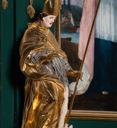 Zdjęcie nr 7: Pełnoplastyczna rzeźba drewniana przedstawiająca św. Stanisława, przyścienna, polichromowana, złocona i srebrzona, wydrążona z tyłu. Ustawiona na niskiej prostokątnej podstawie. Święty ukazany całopostaciowo, w pozycji stojącej w kontrapoście, z prawą nogą ugiętą w kolanie i odstawioną w bok, z wyraźnym przegięciem ciała w lewo i skręceniem korpusu w prawo. W lewej ręce pastorał trzymany na wysokości bioder, prawa zgięta w łokciu i uniesiona przed siebie. Głowa mała, osadzona na długiej szyi, pochylona w dół, lekko zwrócona w lewo. Twarz owalna, policzki pełne. Oczy prawie całkowicie przesłonięte powiekami, wzrok skierowany w dół. Nos duży, prosty, usta małe, pełne, o uniesionych w górę kącikach. Głowę po bokach okalają krótkie włosy układające się w wywinięte na zewnątrz pukle. Na głowie infuła ozdobiona krzyżem równoramiennym o lekko rozszerzających się ramionach, ze wstęgami na plecach. Święty ubrany w albę, rokietę z szeroką, dekoracyjną koronką i mucet. Na ramiona narzucona kapa z galonem ozdobionym stylizowanym motywem roślinnym. Na piersi widoczny pektorał, na szyi humerał, na dłoniach rękawiczki. Spod szat widoczny lewy but. Szaty świętego ukształtowane ekspresyjnie, o ostro ciętych, skośnych fałdach. Poły płaszcza wywinięte na zewnątrz. Odwinięta poła płaszcza pod prawą ręką tworzy obszerny fałd o kształcie litery "S". Z przodu figury, u stóp świętego postać wyłaniającego się z grobu Piotrowina, z mocno odchyloną do tyłu głową nakrytą całunem i wyprostowaną, wyciągniętą pionowo w górę ręką, z widocznie zaznaczonymi mięśniami i dłonią o długich, szczupłych palcach. Twarz Piotrowina wychudzona, z dużym, lekko garbatym nosem, ponad ustami proste wąsy.  Cały strój świętego, z wyjątkiem srebrzonej rokiety, złocony. Twarz i włosy polichromowane. Postać Piotrowina, z wyjątkiem srebrzonego całunu, polichromowana.