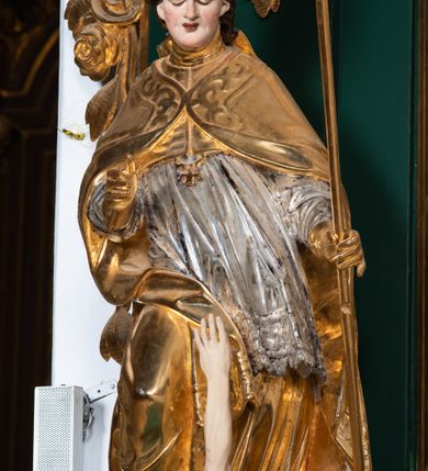 Zdjęcie nr 6: Pełnoplastyczna rzeźba drewniana przedstawiająca św. Stanisława, przyścienna, polichromowana, złocona i srebrzona, wydrążona z tyłu. Ustawiona na niskiej prostokątnej podstawie. Święty ukazany całopostaciowo, w pozycji stojącej w kontrapoście, z prawą nogą ugiętą w kolanie i odstawioną w bok, z wyraźnym przegięciem ciała w lewo i skręceniem korpusu w prawo. W lewej ręce pastorał trzymany na wysokości bioder, prawa zgięta w łokciu i uniesiona przed siebie. Głowa mała, osadzona na długiej szyi, pochylona w dół, lekko zwrócona w lewo. Twarz owalna, policzki pełne. Oczy prawie całkowicie przesłonięte powiekami, wzrok skierowany w dół. Nos duży, prosty, usta małe, pełne, o uniesionych w górę kącikach. Głowę po bokach okalają krótkie włosy układające się w wywinięte na zewnątrz pukle. Na głowie infuła ozdobiona krzyżem równoramiennym o lekko rozszerzających się ramionach, ze wstęgami na plecach. Święty ubrany w albę, rokietę z szeroką, dekoracyjną koronką i mucet. Na ramiona narzucona kapa z galonem ozdobionym stylizowanym motywem roślinnym. Na piersi widoczny pektorał, na szyi humerał, na dłoniach rękawiczki. Spod szat widoczny lewy but. Szaty świętego ukształtowane ekspresyjnie, o ostro ciętych, skośnych fałdach. Poły płaszcza wywinięte na zewnątrz. Odwinięta poła płaszcza pod prawą ręką tworzy obszerny fałd o kształcie litery "S". Z przodu figury, u stóp świętego postać wyłaniającego się z grobu Piotrowina, z mocno odchyloną do tyłu głową nakrytą całunem i wyprostowaną, wyciągniętą pionowo w górę ręką, z widocznie zaznaczonymi mięśniami i dłonią o długich, szczupłych palcach. Twarz Piotrowina wychudzona, z dużym, lekko garbatym nosem, ponad ustami proste wąsy.  Cały strój świętego, z wyjątkiem srebrzonej rokiety, złocony. Twarz i włosy polichromowane. Postać Piotrowina, z wyjątkiem srebrzonego całunu, polichromowana.