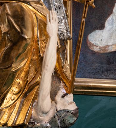 Zdjęcie nr 9: Pełnoplastyczna rzeźba drewniana przedstawiająca św. Stanisława, przyścienna, polichromowana, złocona i srebrzona, wydrążona z tyłu. Ustawiona na niskiej prostokątnej podstawie. Święty ukazany całopostaciowo, w pozycji stojącej w kontrapoście, z prawą nogą ugiętą w kolanie i odstawioną w bok, z wyraźnym przegięciem ciała w lewo i skręceniem korpusu w prawo. W lewej ręce pastorał trzymany na wysokości bioder, prawa zgięta w łokciu i uniesiona przed siebie. Głowa mała, osadzona na długiej szyi, pochylona w dół, lekko zwrócona w lewo. Twarz owalna, policzki pełne. Oczy prawie całkowicie przesłonięte powiekami, wzrok skierowany w dół. Nos duży, prosty, usta małe, pełne, o uniesionych w górę kącikach. Głowę po bokach okalają krótkie włosy układające się w wywinięte na zewnątrz pukle. Na głowie infuła ozdobiona krzyżem równoramiennym o lekko rozszerzających się ramionach, ze wstęgami na plecach. Święty ubrany w albę, rokietę z szeroką, dekoracyjną koronką i mucet. Na ramiona narzucona kapa z galonem ozdobionym stylizowanym motywem roślinnym. Na piersi widoczny pektorał, na szyi humerał, na dłoniach rękawiczki. Spod szat widoczny lewy but. Szaty świętego ukształtowane ekspresyjnie, o ostro ciętych, skośnych fałdach. Poły płaszcza wywinięte na zewnątrz. Odwinięta poła płaszcza pod prawą ręką tworzy obszerny fałd o kształcie litery "S". Z przodu figury, u stóp świętego postać wyłaniającego się z grobu Piotrowina, z mocno odchyloną do tyłu głową nakrytą całunem i wyprostowaną, wyciągniętą pionowo w górę ręką, z widocznie zaznaczonymi mięśniami i dłonią o długich, szczupłych palcach. Twarz Piotrowina wychudzona, z dużym, lekko garbatym nosem, ponad ustami proste wąsy.  Cały strój świętego, z wyjątkiem srebrzonej rokiety, złocony. Twarz i włosy polichromowane. Postać Piotrowina, z wyjątkiem srebrzonego całunu, polichromowana.