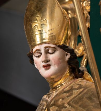 Zdjęcie nr 12: Pełnoplastyczna rzeźba drewniana przedstawiająca św. Stanisława, przyścienna, polichromowana, złocona i srebrzona, wydrążona z tyłu. Ustawiona na niskiej prostokątnej podstawie. Święty ukazany całopostaciowo, w pozycji stojącej w kontrapoście, z prawą nogą ugiętą w kolanie i odstawioną w bok, z wyraźnym przegięciem ciała w lewo i skręceniem korpusu w prawo. W lewej ręce pastorał trzymany na wysokości bioder, prawa zgięta w łokciu i uniesiona przed siebie. Głowa mała, osadzona na długiej szyi, pochylona w dół, lekko zwrócona w lewo. Twarz owalna, policzki pełne. Oczy prawie całkowicie przesłonięte powiekami, wzrok skierowany w dół. Nos duży, prosty, usta małe, pełne, o uniesionych w górę kącikach. Głowę po bokach okalają krótkie włosy układające się w wywinięte na zewnątrz pukle. Na głowie infuła ozdobiona krzyżem równoramiennym o lekko rozszerzających się ramionach, ze wstęgami na plecach. Święty ubrany w albę, rokietę z szeroką, dekoracyjną koronką i mucet. Na ramiona narzucona kapa z galonem ozdobionym stylizowanym motywem roślinnym. Na piersi widoczny pektorał, na szyi humerał, na dłoniach rękawiczki. Spod szat widoczny lewy but. Szaty świętego ukształtowane ekspresyjnie, o ostro ciętych, skośnych fałdach. Poły płaszcza wywinięte na zewnątrz. Odwinięta poła płaszcza pod prawą ręką tworzy obszerny fałd o kształcie litery "S". Z przodu figury, u stóp świętego postać wyłaniającego się z grobu Piotrowina, z mocno odchyloną do tyłu głową nakrytą całunem i wyprostowaną, wyciągniętą pionowo w górę ręką, z widocznie zaznaczonymi mięśniami i dłonią o długich, szczupłych palcach. Twarz Piotrowina wychudzona, z dużym, lekko garbatym nosem, ponad ustami proste wąsy.  Cały strój świętego, z wyjątkiem srebrzonej rokiety, złocony. Twarz i włosy polichromowane. Postać Piotrowina, z wyjątkiem srebrzonego całunu, polichromowana.