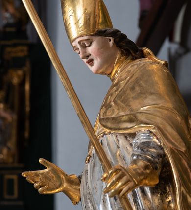 Zdjęcie nr 10: Pełnoplastyczna rzeźba drewniana przedstawiająca św. Stanisława, przyścienna, polichromowana, złocona i srebrzona, wydrążona z tyłu. Ustawiona na niskiej prostokątnej podstawie. Święty ukazany całopostaciowo, w pozycji stojącej w kontrapoście, z prawą nogą ugiętą w kolanie i odstawioną w bok, z wyraźnym przegięciem ciała w lewo i skręceniem korpusu w prawo. W lewej ręce pastorał trzymany na wysokości bioder, prawa zgięta w łokciu i uniesiona przed siebie. Głowa mała, osadzona na długiej szyi, pochylona w dół, lekko zwrócona w lewo. Twarz owalna, policzki pełne. Oczy prawie całkowicie przesłonięte powiekami, wzrok skierowany w dół. Nos duży, prosty, usta małe, pełne, o uniesionych w górę kącikach. Głowę po bokach okalają krótkie włosy układające się w wywinięte na zewnątrz pukle. Na głowie infuła ozdobiona krzyżem równoramiennym o lekko rozszerzających się ramionach, ze wstęgami na plecach. Święty ubrany w albę, rokietę z szeroką, dekoracyjną koronką i mucet. Na ramiona narzucona kapa z galonem ozdobionym stylizowanym motywem roślinnym. Na piersi widoczny pektorał, na szyi humerał, na dłoniach rękawiczki. Spod szat widoczny lewy but. Szaty świętego ukształtowane ekspresyjnie, o ostro ciętych, skośnych fałdach. Poły płaszcza wywinięte na zewnątrz. Odwinięta poła płaszcza pod prawą ręką tworzy obszerny fałd o kształcie litery "S". Z przodu figury, u stóp świętego postać wyłaniającego się z grobu Piotrowina, z mocno odchyloną do tyłu głową nakrytą całunem i wyprostowaną, wyciągniętą pionowo w górę ręką, z widocznie zaznaczonymi mięśniami i dłonią o długich, szczupłych palcach. Twarz Piotrowina wychudzona, z dużym, lekko garbatym nosem, ponad ustami proste wąsy.  Cały strój świętego, z wyjątkiem srebrzonej rokiety, złocony. Twarz i włosy polichromowane. Postać Piotrowina, z wyjątkiem srebrzonego całunu, polichromowana.