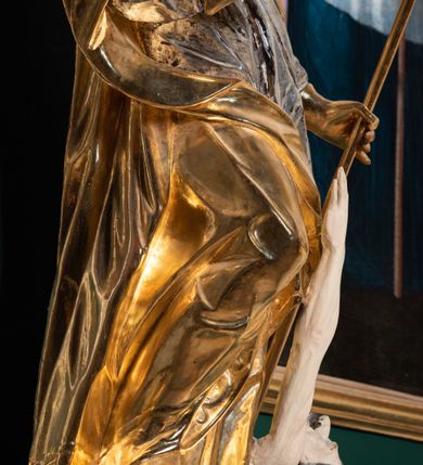 Zdjęcie nr 13: Pełnoplastyczna rzeźba drewniana przedstawiająca św. Stanisława, przyścienna, polichromowana, złocona i srebrzona, wydrążona z tyłu. Ustawiona na niskiej prostokątnej podstawie. Święty ukazany całopostaciowo, w pozycji stojącej w kontrapoście, z prawą nogą ugiętą w kolanie i odstawioną w bok, z wyraźnym przegięciem ciała w lewo i skręceniem korpusu w prawo. W lewej ręce pastorał trzymany na wysokości bioder, prawa zgięta w łokciu i uniesiona przed siebie. Głowa mała, osadzona na długiej szyi, pochylona w dół, lekko zwrócona w lewo. Twarz owalna, policzki pełne. Oczy prawie całkowicie przesłonięte powiekami, wzrok skierowany w dół. Nos duży, prosty, usta małe, pełne, o uniesionych w górę kącikach. Głowę po bokach okalają krótkie włosy układające się w wywinięte na zewnątrz pukle. Na głowie infuła ozdobiona krzyżem równoramiennym o lekko rozszerzających się ramionach, ze wstęgami na plecach. Święty ubrany w albę, rokietę z szeroką, dekoracyjną koronką i mucet. Na ramiona narzucona kapa z galonem ozdobionym stylizowanym motywem roślinnym. Na piersi widoczny pektorał, na szyi humerał, na dłoniach rękawiczki. Spod szat widoczny lewy but. Szaty świętego ukształtowane ekspresyjnie, o ostro ciętych, skośnych fałdach. Poły płaszcza wywinięte na zewnątrz. Odwinięta poła płaszcza pod prawą ręką tworzy obszerny fałd o kształcie litery &quot;S&quot;. Z przodu figury, u stóp świętego postać wyłaniającego się z grobu Piotrowina, z mocno odchyloną do tyłu głową nakrytą całunem i wyprostowaną, wyciągniętą pionowo w górę ręką, z widocznie zaznaczonymi mięśniami i dłonią o długich, szczupłych palcach. Twarz Piotrowina wychudzona, z dużym, lekko garbatym nosem, ponad ustami proste wąsy.  Cały strój świętego, z wyjątkiem srebrzonej rokiety, złocony. Twarz i włosy polichromowane. Postać Piotrowina, z wyjątkiem srebrzonego całunu, polichromowana.