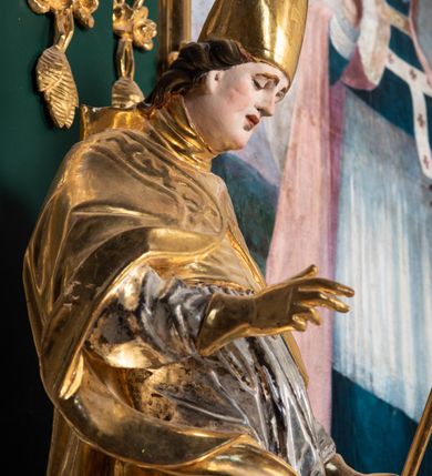 Zdjęcie nr 14: Pełnoplastyczna rzeźba drewniana przedstawiająca św. Stanisława, przyścienna, polichromowana, złocona i srebrzona, wydrążona z tyłu. Ustawiona na niskiej prostokątnej podstawie. Święty ukazany całopostaciowo, w pozycji stojącej w kontrapoście, z prawą nogą ugiętą w kolanie i odstawioną w bok, z wyraźnym przegięciem ciała w lewo i skręceniem korpusu w prawo. W lewej ręce pastorał trzymany na wysokości bioder, prawa zgięta w łokciu i uniesiona przed siebie. Głowa mała, osadzona na długiej szyi, pochylona w dół, lekko zwrócona w lewo. Twarz owalna, policzki pełne. Oczy prawie całkowicie przesłonięte powiekami, wzrok skierowany w dół. Nos duży, prosty, usta małe, pełne, o uniesionych w górę kącikach. Głowę po bokach okalają krótkie włosy układające się w wywinięte na zewnątrz pukle. Na głowie infuła ozdobiona krzyżem równoramiennym o lekko rozszerzających się ramionach, ze wstęgami na plecach. Święty ubrany w albę, rokietę z szeroką, dekoracyjną koronką i mucet. Na ramiona narzucona kapa z galonem ozdobionym stylizowanym motywem roślinnym. Na piersi widoczny pektorał, na szyi humerał, na dłoniach rękawiczki. Spod szat widoczny lewy but. Szaty świętego ukształtowane ekspresyjnie, o ostro ciętych, skośnych fałdach. Poły płaszcza wywinięte na zewnątrz. Odwinięta poła płaszcza pod prawą ręką tworzy obszerny fałd o kształcie litery "S". Z przodu figury, u stóp świętego postać wyłaniającego się z grobu Piotrowina, z mocno odchyloną do tyłu głową nakrytą całunem i wyprostowaną, wyciągniętą pionowo w górę ręką, z widocznie zaznaczonymi mięśniami i dłonią o długich, szczupłych palcach. Twarz Piotrowina wychudzona, z dużym, lekko garbatym nosem, ponad ustami proste wąsy.  Cały strój świętego, z wyjątkiem srebrzonej rokiety, złocony. Twarz i włosy polichromowane. Postać Piotrowina, z wyjątkiem srebrzonego całunu, polichromowana.
