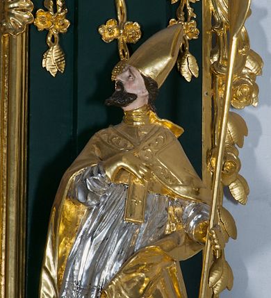 Zdjęcie nr 1: Pełnoplastyczna rzeźba drewniana przedstawiająca św. Wojciecha, przyścienna, polichromowana, złocona i srebrzona, wydrążona z tyłu. Ustawiona na niskiej prostokątnej podstawie. Święty ukazany całopostaciowo, w pozycji stojącej w kontrapoście, z lewą nogą ugiętą w kolanie i odstawioną w bok, z wyraźnym przegięciem ciała w prawo i skręceniem korpusu w lewo. Prawa ręka zgięta w łokciu i nadgarstku spoczywa na piersi, lewa ręka odwiedziona od ciała, ugięta w łokciu i wyciągnięta do przodu, na wysokości pasa trzyma wysokie wiosło. Głowa mała, osadzona na długiej szyi, uniesiona w górę, zwrócona w prawo. Twarz trójkątna, szczupła, podbródek okolony brodą z pofalowanymi i skręconymi pasmami włosów układającymi się w szpic. Wzniesione wyraźnie brwi podkreślają kierunek spojrzenia, nos duży, spiczasty, usta lekko rozchylone, okolone długimi wąsami. Głowę po bokach okalają krótkie włosy, na głowie infuła, ze wstęgą uniesioną nad ramieniem. Święty ubrany jest w albę, rokietę z szeroką, dekoracyjną koronką i mucet. Na ramiona narzucona kapa z galonem ozdobionym stylizowanym motywem roślinnym. Na piersi spod kapy widoczny paliusz, na szyi humerał, na dłoniach rękawiczki. Spod szaty wystają buty. Szaty świętego ukształtowane ekspresyjnie, o ostro ciętych, skośnych fałdach. Poły płaszcza wywinięte na zewnątrz. Odwinięta poła płaszcza pod lewą ręką mocno uniesiona. Cały strój świętego, z wyjątkiem srebrzonej rokiety, złocony. Twarz i włosy polichromowane.