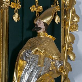 Zdjęcie nr 1: Pełnoplastyczna rzeźba drewniana przedstawiająca św. Wojciecha, przyścienna, polichromowana, złocona i srebrzona, wydrążona z tyłu. Ustawiona na niskiej prostokątnej podstawie. Święty ukazany całopostaciowo, w pozycji stojącej w kontrapoście, z lewą nogą ugiętą w kolanie i odstawioną w bok, z wyraźnym przegięciem ciała w prawo i skręceniem korpusu w lewo. Prawa ręka zgięta w łokciu i nadgarstku spoczywa na piersi, lewa ręka odwiedziona od ciała, ugięta w łokciu i wyciągnięta do przodu, na wysokości pasa trzyma wysokie wiosło. Głowa mała, osadzona na długiej szyi, uniesiona w górę, zwrócona w prawo. Twarz trójkątna, szczupła, podbródek okolony brodą z pofalowanymi i skręconymi pasmami włosów układającymi się w szpic. Wzniesione wyraźnie brwi podkreślają kierunek spojrzenia, nos duży, spiczasty, usta lekko rozchylone, okolone długimi wąsami. Głowę po bokach okalają krótkie włosy, na głowie infuła, ze wstęgą uniesioną nad ramieniem. Święty ubrany jest w albę, rokietę z szeroką, dekoracyjną koronką i mucet. Na ramiona narzucona kapa z galonem ozdobionym stylizowanym motywem roślinnym. Na piersi spod kapy widoczny paliusz, na szyi humerał, na dłoniach rękawiczki. Spod szaty wystają buty. Szaty świętego ukształtowane ekspresyjnie, o ostro ciętych, skośnych fałdach. Poły płaszcza wywinięte na zewnątrz. Odwinięta poła płaszcza pod lewą ręką mocno uniesiona. Cały strój świętego, z wyjątkiem srebrzonej rokiety, złocony. Twarz i włosy polichromowane.