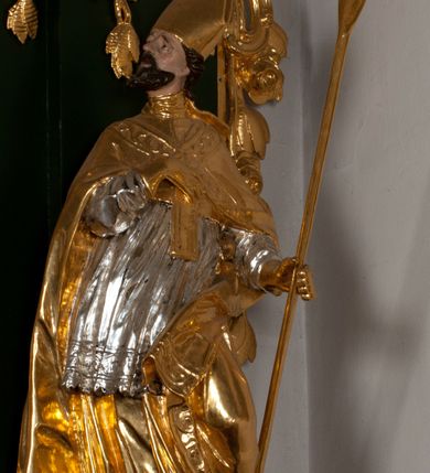 Zdjęcie nr 8: Pełnoplastyczna rzeźba drewniana przedstawiająca św. Wojciecha, przyścienna, polichromowana, złocona i srebrzona, wydrążona z tyłu. Ustawiona na niskiej prostokątnej podstawie. Święty ukazany całopostaciowo, w pozycji stojącej w kontrapoście, z lewą nogą ugiętą w kolanie i odstawioną w bok, z wyraźnym przegięciem ciała w prawo i skręceniem korpusu w lewo. Prawa ręka zgięta w łokciu i nadgarstku spoczywa na piersi, lewa ręka odwiedziona od ciała, ugięta w łokciu i wyciągnięta do przodu, na wysokości pasa trzyma wysokie wiosło. Głowa mała, osadzona na długiej szyi, uniesiona w górę, zwrócona w prawo. Twarz trójkątna, szczupła, podbródek okolony brodą z pofalowanymi i skręconymi pasmami włosów układającymi się w szpic. Wzniesione wyraźnie brwi podkreślają kierunek spojrzenia, nos duży, spiczasty, usta lekko rozchylone, okolone długimi wąsami. Głowę po bokach okalają krótkie włosy, na głowie infuła, ze wstęgą uniesioną nad ramieniem. Święty ubrany jest w albę, rokietę z szeroką, dekoracyjną koronką i mucet. Na ramiona narzucona kapa z galonem ozdobionym stylizowanym motywem roślinnym. Na piersi spod kapy widoczny paliusz, na szyi humerał, na dłoniach rękawiczki. Spod szaty wystają buty. Szaty świętego ukształtowane ekspresyjnie, o ostro ciętych, skośnych fałdach. Poły płaszcza wywinięte na zewnątrz. Odwinięta poła płaszcza pod lewą ręką mocno uniesiona. Cały strój świętego, z wyjątkiem srebrzonej rokiety, złocony. Twarz i włosy polichromowane.