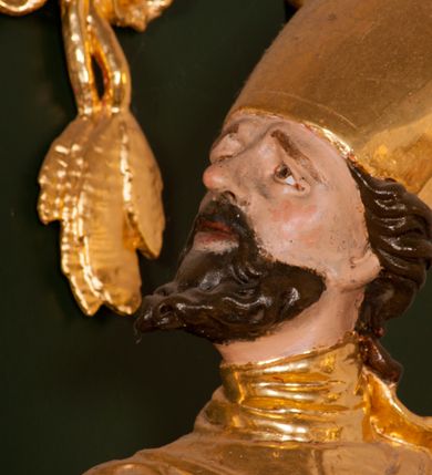 Zdjęcie nr 11: Pełnoplastyczna rzeźba drewniana przedstawiająca św. Wojciecha, przyścienna, polichromowana, złocona i srebrzona, wydrążona z tyłu. Ustawiona na niskiej prostokątnej podstawie. Święty ukazany całopostaciowo, w pozycji stojącej w kontrapoście, z lewą nogą ugiętą w kolanie i odstawioną w bok, z wyraźnym przegięciem ciała w prawo i skręceniem korpusu w lewo. Prawa ręka zgięta w łokciu i nadgarstku spoczywa na piersi, lewa ręka odwiedziona od ciała, ugięta w łokciu i wyciągnięta do przodu, na wysokości pasa trzyma wysokie wiosło. Głowa mała, osadzona na długiej szyi, uniesiona w górę, zwrócona w prawo. Twarz trójkątna, szczupła, podbródek okolony brodą z pofalowanymi i skręconymi pasmami włosów układającymi się w szpic. Wzniesione wyraźnie brwi podkreślają kierunek spojrzenia, nos duży, spiczasty, usta lekko rozchylone, okolone długimi wąsami. Głowę po bokach okalają krótkie włosy, na głowie infuła, ze wstęgą uniesioną nad ramieniem. Święty ubrany jest w albę, rokietę z szeroką, dekoracyjną koronką i mucet. Na ramiona narzucona kapa z galonem ozdobionym stylizowanym motywem roślinnym. Na piersi spod kapy widoczny paliusz, na szyi humerał, na dłoniach rękawiczki. Spod szaty wystają buty. Szaty świętego ukształtowane ekspresyjnie, o ostro ciętych, skośnych fałdach. Poły płaszcza wywinięte na zewnątrz. Odwinięta poła płaszcza pod lewą ręką mocno uniesiona. Cały strój świętego, z wyjątkiem srebrzonej rokiety, złocony. Twarz i włosy polichromowane.