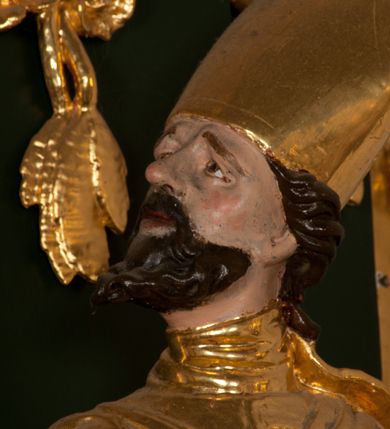 Zdjęcie nr 9: Pełnoplastyczna rzeźba drewniana przedstawiająca św. Wojciecha, przyścienna, polichromowana, złocona i srebrzona, wydrążona z tyłu. Ustawiona na niskiej prostokątnej podstawie. Święty ukazany całopostaciowo, w pozycji stojącej w kontrapoście, z lewą nogą ugiętą w kolanie i odstawioną w bok, z wyraźnym przegięciem ciała w prawo i skręceniem korpusu w lewo. Prawa ręka zgięta w łokciu i nadgarstku spoczywa na piersi, lewa ręka odwiedziona od ciała, ugięta w łokciu i wyciągnięta do przodu, na wysokości pasa trzyma wysokie wiosło. Głowa mała, osadzona na długiej szyi, uniesiona w górę, zwrócona w prawo. Twarz trójkątna, szczupła, podbródek okolony brodą z pofalowanymi i skręconymi pasmami włosów układającymi się w szpic. Wzniesione wyraźnie brwi podkreślają kierunek spojrzenia, nos duży, spiczasty, usta lekko rozchylone, okolone długimi wąsami. Głowę po bokach okalają krótkie włosy, na głowie infuła, ze wstęgą uniesioną nad ramieniem. Święty ubrany jest w albę, rokietę z szeroką, dekoracyjną koronką i mucet. Na ramiona narzucona kapa z galonem ozdobionym stylizowanym motywem roślinnym. Na piersi spod kapy widoczny paliusz, na szyi humerał, na dłoniach rękawiczki. Spod szaty wystają buty. Szaty świętego ukształtowane ekspresyjnie, o ostro ciętych, skośnych fałdach. Poły płaszcza wywinięte na zewnątrz. Odwinięta poła płaszcza pod lewą ręką mocno uniesiona. Cały strój świętego, z wyjątkiem srebrzonej rokiety, złocony. Twarz i włosy polichromowane.