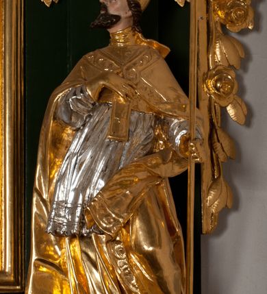 Zdjęcie nr 13: Pełnoplastyczna rzeźba drewniana przedstawiająca św. Wojciecha, przyścienna, polichromowana, złocona i srebrzona, wydrążona z tyłu. Ustawiona na niskiej prostokątnej podstawie. Święty ukazany całopostaciowo, w pozycji stojącej w kontrapoście, z lewą nogą ugiętą w kolanie i odstawioną w bok, z wyraźnym przegięciem ciała w prawo i skręceniem korpusu w lewo. Prawa ręka zgięta w łokciu i nadgarstku spoczywa na piersi, lewa ręka odwiedziona od ciała, ugięta w łokciu i wyciągnięta do przodu, na wysokości pasa trzyma wysokie wiosło. Głowa mała, osadzona na długiej szyi, uniesiona w górę, zwrócona w prawo. Twarz trójkątna, szczupła, podbródek okolony brodą z pofalowanymi i skręconymi pasmami włosów układającymi się w szpic. Wzniesione wyraźnie brwi podkreślają kierunek spojrzenia, nos duży, spiczasty, usta lekko rozchylone, okolone długimi wąsami. Głowę po bokach okalają krótkie włosy, na głowie infuła, ze wstęgą uniesioną nad ramieniem. Święty ubrany jest w albę, rokietę z szeroką, dekoracyjną koronką i mucet. Na ramiona narzucona kapa z galonem ozdobionym stylizowanym motywem roślinnym. Na piersi spod kapy widoczny paliusz, na szyi humerał, na dłoniach rękawiczki. Spod szaty wystają buty. Szaty świętego ukształtowane ekspresyjnie, o ostro ciętych, skośnych fałdach. Poły płaszcza wywinięte na zewnątrz. Odwinięta poła płaszcza pod lewą ręką mocno uniesiona. Cały strój świętego, z wyjątkiem srebrzonej rokiety, złocony. Twarz i włosy polichromowane.