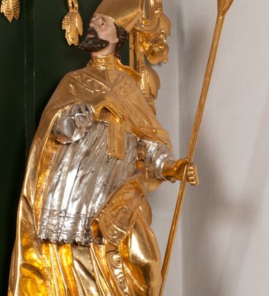 Zdjęcie nr 12: Pełnoplastyczna rzeźba drewniana przedstawiająca św. Wojciecha, przyścienna, polichromowana, złocona i srebrzona, wydrążona z tyłu. Ustawiona na niskiej prostokątnej podstawie. Święty ukazany całopostaciowo, w pozycji stojącej w kontrapoście, z lewą nogą ugiętą w kolanie i odstawioną w bok, z wyraźnym przegięciem ciała w prawo i skręceniem korpusu w lewo. Prawa ręka zgięta w łokciu i nadgarstku spoczywa na piersi, lewa ręka odwiedziona od ciała, ugięta w łokciu i wyciągnięta do przodu, na wysokości pasa trzyma wysokie wiosło. Głowa mała, osadzona na długiej szyi, uniesiona w górę, zwrócona w prawo. Twarz trójkątna, szczupła, podbródek okolony brodą z pofalowanymi i skręconymi pasmami włosów układającymi się w szpic. Wzniesione wyraźnie brwi podkreślają kierunek spojrzenia, nos duży, spiczasty, usta lekko rozchylone, okolone długimi wąsami. Głowę po bokach okalają krótkie włosy, na głowie infuła, ze wstęgą uniesioną nad ramieniem. Święty ubrany jest w albę, rokietę z szeroką, dekoracyjną koronką i mucet. Na ramiona narzucona kapa z galonem ozdobionym stylizowanym motywem roślinnym. Na piersi spod kapy widoczny paliusz, na szyi humerał, na dłoniach rękawiczki. Spod szaty wystają buty. Szaty świętego ukształtowane ekspresyjnie, o ostro ciętych, skośnych fałdach. Poły płaszcza wywinięte na zewnątrz. Odwinięta poła płaszcza pod lewą ręką mocno uniesiona. Cały strój świętego, z wyjątkiem srebrzonej rokiety, złocony. Twarz i włosy polichromowane.