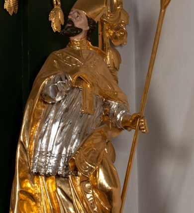 Zdjęcie nr 10: Pełnoplastyczna rzeźba drewniana przedstawiająca św. Wojciecha, przyścienna, polichromowana, złocona i srebrzona, wydrążona z tyłu. Ustawiona na niskiej prostokątnej podstawie. Święty ukazany całopostaciowo, w pozycji stojącej w kontrapoście, z lewą nogą ugiętą w kolanie i odstawioną w bok, z wyraźnym przegięciem ciała w prawo i skręceniem korpusu w lewo. Prawa ręka zgięta w łokciu i nadgarstku spoczywa na piersi, lewa ręka odwiedziona od ciała, ugięta w łokciu i wyciągnięta do przodu, na wysokości pasa trzyma wysokie wiosło. Głowa mała, osadzona na długiej szyi, uniesiona w górę, zwrócona w prawo. Twarz trójkątna, szczupła, podbródek okolony brodą z pofalowanymi i skręconymi pasmami włosów układającymi się w szpic. Wzniesione wyraźnie brwi podkreślają kierunek spojrzenia, nos duży, spiczasty, usta lekko rozchylone, okolone długimi wąsami. Głowę po bokach okalają krótkie włosy, na głowie infuła, ze wstęgą uniesioną nad ramieniem. Święty ubrany jest w albę, rokietę z szeroką, dekoracyjną koronką i mucet. Na ramiona narzucona kapa z galonem ozdobionym stylizowanym motywem roślinnym. Na piersi spod kapy widoczny paliusz, na szyi humerał, na dłoniach rękawiczki. Spod szaty wystają buty. Szaty świętego ukształtowane ekspresyjnie, o ostro ciętych, skośnych fałdach. Poły płaszcza wywinięte na zewnątrz. Odwinięta poła płaszcza pod lewą ręką mocno uniesiona. Cały strój świętego, z wyjątkiem srebrzonej rokiety, złocony. Twarz i włosy polichromowane.