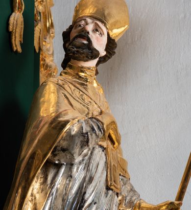 Zdjęcie nr 5: Pełnoplastyczna rzeźba drewniana przedstawiająca św. Wojciecha, przyścienna, polichromowana, złocona i srebrzona, wydrążona z tyłu. Ustawiona na niskiej prostokątnej podstawie. Święty ukazany całopostaciowo, w pozycji stojącej w kontrapoście, z lewą nogą ugiętą w kolanie i odstawioną w bok, z wyraźnym przegięciem ciała w prawo i skręceniem korpusu w lewo. Prawa ręka zgięta w łokciu i nadgarstku spoczywa na piersi, lewa ręka odwiedziona od ciała, ugięta w łokciu i wyciągnięta do przodu, na wysokości pasa trzyma wysokie wiosło. Głowa mała, osadzona na długiej szyi, uniesiona w górę, zwrócona w prawo. Twarz trójkątna, szczupła, podbródek okolony brodą z pofalowanymi i skręconymi pasmami włosów układającymi się w szpic. Wzniesione wyraźnie brwi podkreślają kierunek spojrzenia, nos duży, spiczasty, usta lekko rozchylone, okolone długimi wąsami. Głowę po bokach okalają krótkie włosy, na głowie infuła, ze wstęgą uniesioną nad ramieniem. Święty ubrany jest w albę, rokietę z szeroką, dekoracyjną koronką i mucet. Na ramiona narzucona kapa z galonem ozdobionym stylizowanym motywem roślinnym. Na piersi spod kapy widoczny paliusz, na szyi humerał, na dłoniach rękawiczki. Spod szaty wystają buty. Szaty świętego ukształtowane ekspresyjnie, o ostro ciętych, skośnych fałdach. Poły płaszcza wywinięte na zewnątrz. Odwinięta poła płaszcza pod lewą ręką mocno uniesiona. Cały strój świętego, z wyjątkiem srebrzonej rokiety, złocony. Twarz i włosy polichromowane.