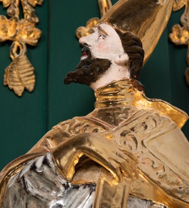 Zdjęcie nr 4: Pełnoplastyczna rzeźba drewniana przedstawiająca św. Wojciecha, przyścienna, polichromowana, złocona i srebrzona, wydrążona z tyłu. Ustawiona na niskiej prostokątnej podstawie. Święty ukazany całopostaciowo, w pozycji stojącej w kontrapoście, z lewą nogą ugiętą w kolanie i odstawioną w bok, z wyraźnym przegięciem ciała w prawo i skręceniem korpusu w lewo. Prawa ręka zgięta w łokciu i nadgarstku spoczywa na piersi, lewa ręka odwiedziona od ciała, ugięta w łokciu i wyciągnięta do przodu, na wysokości pasa trzyma wysokie wiosło. Głowa mała, osadzona na długiej szyi, uniesiona w górę, zwrócona w prawo. Twarz trójkątna, szczupła, podbródek okolony brodą z pofalowanymi i skręconymi pasmami włosów układającymi się w szpic. Wzniesione wyraźnie brwi podkreślają kierunek spojrzenia, nos duży, spiczasty, usta lekko rozchylone, okolone długimi wąsami. Głowę po bokach okalają krótkie włosy, na głowie infuła, ze wstęgą uniesioną nad ramieniem. Święty ubrany jest w albę, rokietę z szeroką, dekoracyjną koronką i mucet. Na ramiona narzucona kapa z galonem ozdobionym stylizowanym motywem roślinnym. Na piersi spod kapy widoczny paliusz, na szyi humerał, na dłoniach rękawiczki. Spod szaty wystają buty. Szaty świętego ukształtowane ekspresyjnie, o ostro ciętych, skośnych fałdach. Poły płaszcza wywinięte na zewnątrz. Odwinięta poła płaszcza pod lewą ręką mocno uniesiona. Cały strój świętego, z wyjątkiem srebrzonej rokiety, złocony. Twarz i włosy polichromowane.