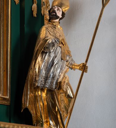 Zdjęcie nr 6: Pełnoplastyczna rzeźba drewniana przedstawiająca św. Wojciecha, przyścienna, polichromowana, złocona i srebrzona, wydrążona z tyłu. Ustawiona na niskiej prostokątnej podstawie. Święty ukazany całopostaciowo, w pozycji stojącej w kontrapoście, z lewą nogą ugiętą w kolanie i odstawioną w bok, z wyraźnym przegięciem ciała w prawo i skręceniem korpusu w lewo. Prawa ręka zgięta w łokciu i nadgarstku spoczywa na piersi, lewa ręka odwiedziona od ciała, ugięta w łokciu i wyciągnięta do przodu, na wysokości pasa trzyma wysokie wiosło. Głowa mała, osadzona na długiej szyi, uniesiona w górę, zwrócona w prawo. Twarz trójkątna, szczupła, podbródek okolony brodą z pofalowanymi i skręconymi pasmami włosów układającymi się w szpic. Wzniesione wyraźnie brwi podkreślają kierunek spojrzenia, nos duży, spiczasty, usta lekko rozchylone, okolone długimi wąsami. Głowę po bokach okalają krótkie włosy, na głowie infuła, ze wstęgą uniesioną nad ramieniem. Święty ubrany jest w albę, rokietę z szeroką, dekoracyjną koronką i mucet. Na ramiona narzucona kapa z galonem ozdobionym stylizowanym motywem roślinnym. Na piersi spod kapy widoczny paliusz, na szyi humerał, na dłoniach rękawiczki. Spod szaty wystają buty. Szaty świętego ukształtowane ekspresyjnie, o ostro ciętych, skośnych fałdach. Poły płaszcza wywinięte na zewnątrz. Odwinięta poła płaszcza pod lewą ręką mocno uniesiona. Cały strój świętego, z wyjątkiem srebrzonej rokiety, złocony. Twarz i włosy polichromowane.