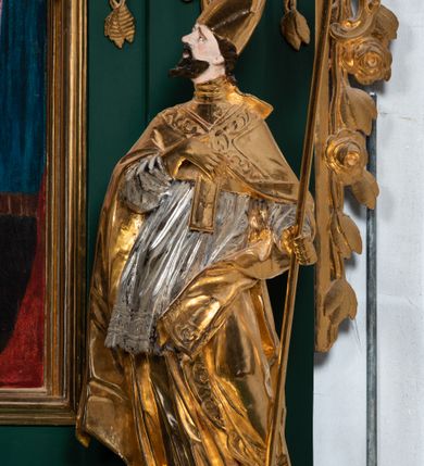 Zdjęcie nr 7: Pełnoplastyczna rzeźba drewniana przedstawiająca św. Wojciecha, przyścienna, polichromowana, złocona i srebrzona, wydrążona z tyłu. Ustawiona na niskiej prostokątnej podstawie. Święty ukazany całopostaciowo, w pozycji stojącej w kontrapoście, z lewą nogą ugiętą w kolanie i odstawioną w bok, z wyraźnym przegięciem ciała w prawo i skręceniem korpusu w lewo. Prawa ręka zgięta w łokciu i nadgarstku spoczywa na piersi, lewa ręka odwiedziona od ciała, ugięta w łokciu i wyciągnięta do przodu, na wysokości pasa trzyma wysokie wiosło. Głowa mała, osadzona na długiej szyi, uniesiona w górę, zwrócona w prawo. Twarz trójkątna, szczupła, podbródek okolony brodą z pofalowanymi i skręconymi pasmami włosów układającymi się w szpic. Wzniesione wyraźnie brwi podkreślają kierunek spojrzenia, nos duży, spiczasty, usta lekko rozchylone, okolone długimi wąsami. Głowę po bokach okalają krótkie włosy, na głowie infuła, ze wstęgą uniesioną nad ramieniem. Święty ubrany jest w albę, rokietę z szeroką, dekoracyjną koronką i mucet. Na ramiona narzucona kapa z galonem ozdobionym stylizowanym motywem roślinnym. Na piersi spod kapy widoczny paliusz, na szyi humerał, na dłoniach rękawiczki. Spod szaty wystają buty. Szaty świętego ukształtowane ekspresyjnie, o ostro ciętych, skośnych fałdach. Poły płaszcza wywinięte na zewnątrz. Odwinięta poła płaszcza pod lewą ręką mocno uniesiona. Cały strój świętego, z wyjątkiem srebrzonej rokiety, złocony. Twarz i włosy polichromowane.