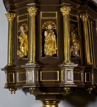 Zdjęcie nr 13: Ambona drewniana, przyścienna, wisząca, dostawiona do północnej ściany nawy ze schodkami biegnącymi wzdłuż ściany. Kosz ambony jest sześcioboczny, z niewielkim parapetem. Jego boki zostały podzielone na pola stojącymi na wysokich cokołach pięcioma kolumienkami o korynckich kapitelach, dźwigających profilowane belkowanie. W polach prostokątne płyciny z konchowymi wnękami ozdobionymi muszlami z figurami ewangelistów. 
We wnękach rzeźby z przedstawieniami ewangelistów i Chrystusa. Figury drewniane pełnoplastyczne, wydrążone od tyłu, polichromowane, złocone i srebrzone, ustawione na niewielkich prostokątnych podstawach, od lewej: św. Łukasz wsparty prawą nogą na głowie byka, św. Marek wsparty prawą nogą na głowie lwa, św. Jan z orłem przy prawej nodze, Chrystus Salvator Mundi, św. Mateusz z aniołem przy prawej nodze. Ewangeliści trzymają w prawych dłoniach otwarte księgi, z wyjątkiem św. Mateusza, trzymającego kodeks w lewej ręce. Dłonie ewangelistów nieproporcjonalnie duże. Szaty bogato pofałdowane, głęboko cięte. 
Podstawa kosza bogato profilowana, ozdobiona podwieszoną dekoracyjną szyszką. Schodki ograniczone parapetem z trzema romboidalnymi płycinami w złoconych profilowanych obramieniach. Na filarze przy wejściu na schodki złocona kula z szyszką. Zaplecek w kształcie stojącego prostokąta, w górnej części ujęty zwężającymi się ku dołowi pilastrami wypełnionymi cekinami, zwieńczonymi rombowymi wypustkami i korynckimi kapitelami, które podtrzymują przełamujące się nad nimi belkowanie. Pośrodku zaplecka w prostokątnym obramieniu zamkniętym nadwieszonym łukiem półkolistym płaskorzeźbione popiersie Boga Ojca wyłaniającego się z obłoków. Prawa ręka Boga wzniesiona w geście błogosławieństwa, lewa oparta na globie ziemskim zwieńczonym krzyżem. Tłem dla postaci Boga Ojca jest rozwiana poła płaszcza układająca się niemal półkoliście. Baldachim ośmioboczny. W profilowanym podniebiu, ozdobionym pasem ornamentu małżowinowo-chrząstkowego i profilowanymi wisiorami, gołębica Ducha Świętego na tle glorii utworzonej z prostych promieni o równej długości. Baldachim ozdobiony zamkniętą koroną, którą tworzy pięć esownic pokrytych pasami ornamentu małżowinowo-chrząstkowego. W zwieńczeniu pełnoplastyczna rzeźba przedstawiająca św. Jana Chrzciciela. Figura drewniana, wydrążona z tyłu, polichromowana, złocona i srebrzona, ustawiona na niewielkiej wielobocznej profilowanej podstawie. Święty w prawej ręce trzyma wysoki krzyż, w lewej księgę, na której spoczywa Baranek Boży. Struktura ambony ciemnobrązowa. Kolumny, kapitele, pilastry w zaplecku korona baldachimu, profilowania i ornamenty złocone, z wyjątkiem srebrzonej ramy w zaplecku i gołębicy Ducha Świętego.