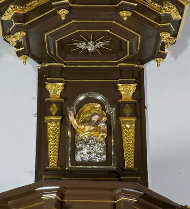 Zdjęcie nr 23: Ambona drewniana, przyścienna, wisząca, dostawiona do północnej ściany nawy ze schodkami biegnącymi wzdłuż ściany. Kosz ambony jest sześcioboczny, z niewielkim parapetem. Jego boki zostały podzielone na pola stojącymi na wysokich cokołach pięcioma kolumienkami o korynckich kapitelach, dźwigających profilowane belkowanie. W polach prostokątne płyciny z konchowymi wnękami ozdobionymi muszlami z figurami ewangelistów. 
We wnękach rzeźby z przedstawieniami ewangelistów i Chrystusa. Figury drewniane pełnoplastyczne, wydrążone od tyłu, polichromowane, złocone i srebrzone, ustawione na niewielkich prostokątnych podstawach, od lewej: św. Łukasz wsparty prawą nogą na głowie byka, św. Marek wsparty prawą nogą na głowie lwa, św. Jan z orłem przy prawej nodze, Chrystus Salvator Mundi, św. Mateusz z aniołem przy prawej nodze. Ewangeliści trzymają w prawych dłoniach otwarte księgi, z wyjątkiem św. Mateusza, trzymającego kodeks w lewej ręce. Dłonie ewangelistów nieproporcjonalnie duże. Szaty bogato pofałdowane, głęboko cięte. 
Podstawa kosza bogato profilowana, ozdobiona podwieszoną dekoracyjną szyszką. Schodki ograniczone parapetem z trzema romboidalnymi płycinami w złoconych profilowanych obramieniach. Na filarze przy wejściu na schodki złocona kula z szyszką. Zaplecek w kształcie stojącego prostokąta, w górnej części ujęty zwężającymi się ku dołowi pilastrami wypełnionymi cekinami, zwieńczonymi rombowymi wypustkami i korynckimi kapitelami, które podtrzymują przełamujące się nad nimi belkowanie. Pośrodku zaplecka w prostokątnym obramieniu zamkniętym nadwieszonym łukiem półkolistym płaskorzeźbione popiersie Boga Ojca wyłaniającego się z obłoków. Prawa ręka Boga wzniesiona w geście błogosławieństwa, lewa oparta na globie ziemskim zwieńczonym krzyżem. Tłem dla postaci Boga Ojca jest rozwiana poła płaszcza układająca się niemal półkoliście. Baldachim ośmioboczny. W profilowanym podniebiu, ozdobionym pasem ornamentu małżowinowo-chrząstkowego i profilowanymi wisiorami, gołębica Ducha Świętego na tle glorii utworzonej z prostych promieni o równej długości. Baldachim ozdobiony zamkniętą koroną, którą tworzy pięć esownic pokrytych pasami ornamentu małżowinowo-chrząstkowego. W zwieńczeniu pełnoplastyczna rzeźba przedstawiająca św. Jana Chrzciciela. Figura drewniana, wydrążona z tyłu, polichromowana, złocona i srebrzona, ustawiona na niewielkiej wielobocznej profilowanej podstawie. Święty w prawej ręce trzyma wysoki krzyż, w lewej księgę, na której spoczywa Baranek Boży. Struktura ambony ciemnobrązowa. Kolumny, kapitele, pilastry w zaplecku korona baldachimu, profilowania i ornamenty złocone, z wyjątkiem srebrzonej ramy w zaplecku i gołębicy Ducha Świętego.