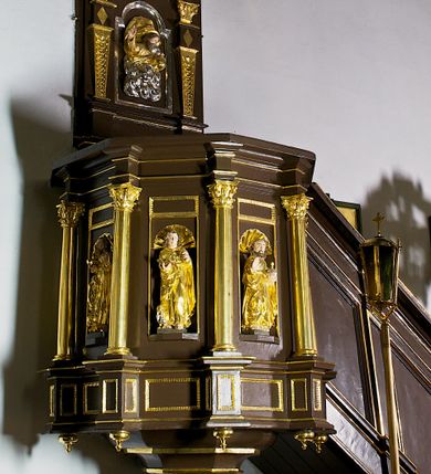 Zdjęcie nr 1: Ambona drewniana, przyścienna, wisząca, dostawiona do północnej ściany nawy ze schodkami biegnącymi wzdłuż ściany. Kosz ambony jest sześcioboczny, z niewielkim parapetem. Jego boki zostały podzielone na pola stojącymi na wysokich cokołach pięcioma kolumienkami o korynckich kapitelach, dźwigających profilowane belkowanie. W polach prostokątne płyciny z konchowymi wnękami ozdobionymi muszlami z figurami ewangelistów. 
We wnękach rzeźby z przedstawieniami ewangelistów i Chrystusa. Figury drewniane pełnoplastyczne, wydrążone od tyłu, polichromowane, złocone i srebrzone, ustawione na niewielkich prostokątnych podstawach, od lewej: św. Łukasz wsparty prawą nogą na głowie byka, św. Marek wsparty prawą nogą na głowie lwa, św. Jan z orłem przy prawej nodze, Chrystus Salvator Mundi, św. Mateusz z aniołem przy prawej nodze. Ewangeliści trzymają w prawych dłoniach otwarte księgi, z wyjątkiem św. Mateusza, trzymającego kodeks w lewej ręce. Dłonie ewangelistów nieproporcjonalnie duże. Szaty bogato pofałdowane, głęboko cięte. 
Podstawa kosza bogato profilowana, ozdobiona podwieszoną dekoracyjną szyszką. Schodki ograniczone parapetem z trzema romboidalnymi płycinami w złoconych profilowanych obramieniach. Na filarze przy wejściu na schodki złocona kula z szyszką. Zaplecek w kształcie stojącego prostokąta, w górnej części ujęty zwężającymi się ku dołowi pilastrami wypełnionymi cekinami, zwieńczonymi rombowymi wypustkami i korynckimi kapitelami, które podtrzymują przełamujące się nad nimi belkowanie. Pośrodku zaplecka w prostokątnym obramieniu zamkniętym nadwieszonym łukiem półkolistym płaskorzeźbione popiersie Boga Ojca wyłaniającego się z obłoków. Prawa ręka Boga wzniesiona w geście błogosławieństwa, lewa oparta na globie ziemskim zwieńczonym krzyżem. Tłem dla postaci Boga Ojca jest rozwiana poła płaszcza układająca się niemal półkoliście. Baldachim ośmioboczny. W profilowanym podniebiu, ozdobionym pasem ornamentu małżowinowo-chrząstkowego i profilowanymi wisiorami, gołębica Ducha Świętego na tle glorii utworzonej z prostych promieni o równej długości. Baldachim ozdobiony zamkniętą koroną, którą tworzy pięć esownic pokrytych pasami ornamentu małżowinowo-chrząstkowego. W zwieńczeniu pełnoplastyczna rzeźba przedstawiająca św. Jana Chrzciciela. Figura drewniana, wydrążona z tyłu, polichromowana, złocona i srebrzona, ustawiona na niewielkiej wielobocznej profilowanej podstawie. Święty w prawej ręce trzyma wysoki krzyż, w lewej księgę, na której spoczywa Baranek Boży. Struktura ambony ciemnobrązowa. Kolumny, kapitele, pilastry w zaplecku korona baldachimu, profilowania i ornamenty złocone, z wyjątkiem srebrzonej ramy w zaplecku i gołębicy Ducha Świętego.