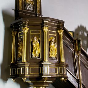Zdjęcie nr 1: Ambona drewniana, przyścienna, wisząca, dostawiona do północnej ściany nawy ze schodkami biegnącymi wzdłuż ściany. Kosz ambony jest sześcioboczny, z niewielkim parapetem. Jego boki zostały podzielone na pola stojącymi na wysokich cokołach pięcioma kolumienkami o korynckich kapitelach, dźwigających profilowane belkowanie. W polach prostokątne płyciny z konchowymi wnękami ozdobionymi muszlami z figurami ewangelistów. 
We wnękach rzeźby z przedstawieniami ewangelistów i Chrystusa. Figury drewniane pełnoplastyczne, wydrążone od tyłu, polichromowane, złocone i srebrzone, ustawione na niewielkich prostokątnych podstawach, od lewej: św. Łukasz wsparty prawą nogą na głowie byka, św. Marek wsparty prawą nogą na głowie lwa, św. Jan z orłem przy prawej nodze, Chrystus Salvator Mundi, św. Mateusz z aniołem przy prawej nodze. Ewangeliści trzymają w prawych dłoniach otwarte księgi, z wyjątkiem św. Mateusza, trzymającego kodeks w lewej ręce. Dłonie ewangelistów nieproporcjonalnie duże. Szaty bogato pofałdowane, głęboko cięte. 
Podstawa kosza bogato profilowana, ozdobiona podwieszoną dekoracyjną szyszką. Schodki ograniczone parapetem z trzema romboidalnymi płycinami w złoconych profilowanych obramieniach. Na filarze przy wejściu na schodki złocona kula z szyszką. Zaplecek w kształcie stojącego prostokąta, w górnej części ujęty zwężającymi się ku dołowi pilastrami wypełnionymi cekinami, zwieńczonymi rombowymi wypustkami i korynckimi kapitelami, które podtrzymują przełamujące się nad nimi belkowanie. Pośrodku zaplecka w prostokątnym obramieniu zamkniętym nadwieszonym łukiem półkolistym płaskorzeźbione popiersie Boga Ojca wyłaniającego się z obłoków. Prawa ręka Boga wzniesiona w geście błogosławieństwa, lewa oparta na globie ziemskim zwieńczonym krzyżem. Tłem dla postaci Boga Ojca jest rozwiana poła płaszcza układająca się niemal półkoliście. Baldachim ośmioboczny. W profilowanym podniebiu, ozdobionym pasem ornamentu małżowinowo-chrząstkowego i profilowanymi wisiorami, gołębica Ducha Świętego na tle glorii utworzonej z prostych promieni o równej długości. Baldachim ozdobiony zamkniętą koroną, którą tworzy pięć esownic pokrytych pasami ornamentu małżowinowo-chrząstkowego. W zwieńczeniu pełnoplastyczna rzeźba przedstawiająca św. Jana Chrzciciela. Figura drewniana, wydrążona z tyłu, polichromowana, złocona i srebrzona, ustawiona na niewielkiej wielobocznej profilowanej podstawie. Święty w prawej ręce trzyma wysoki krzyż, w lewej księgę, na której spoczywa Baranek Boży. Struktura ambony ciemnobrązowa. Kolumny, kapitele, pilastry w zaplecku korona baldachimu, profilowania i ornamenty złocone, z wyjątkiem srebrzonej ramy w zaplecku i gołębicy Ducha Świętego.