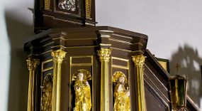 Zdjęcie nr 1: Ambona drewniana, przyścienna, wisząca, dostawiona do północnej ściany nawy ze schodkami biegnącymi wzdłuż ściany. Kosz ambony jest sześcioboczny, z niewielkim parapetem. Jego boki zostały podzielone na pola stojącymi na wysokich cokołach pięcioma kolumienkami o korynckich kapitelach, dźwigających profilowane belkowanie. W polach prostokątne płyciny z konchowymi wnękami ozdobionymi muszlami z figurami ewangelistów. 
We wnękach rzeźby z przedstawieniami ewangelistów i Chrystusa. Figury drewniane pełnoplastyczne, wydrążone od tyłu, polichromowane, złocone i srebrzone, ustawione na niewielkich prostokątnych podstawach, od lewej: św. Łukasz wsparty prawą nogą na głowie byka, św. Marek wsparty prawą nogą na głowie lwa, św. Jan z orłem przy prawej nodze, Chrystus Salvator Mundi, św. Mateusz z aniołem przy prawej nodze. Ewangeliści trzymają w prawych dłoniach otwarte księgi, z wyjątkiem św. Mateusza, trzymającego kodeks w lewej ręce. Dłonie ewangelistów nieproporcjonalnie duże. Szaty bogato pofałdowane, głęboko cięte. 
Podstawa kosza bogato profilowana, ozdobiona podwieszoną dekoracyjną szyszką. Schodki ograniczone parapetem z trzema romboidalnymi płycinami w złoconych profilowanych obramieniach. Na filarze przy wejściu na schodki złocona kula z szyszką. Zaplecek w kształcie stojącego prostokąta, w górnej części ujęty zwężającymi się ku dołowi pilastrami wypełnionymi cekinami, zwieńczonymi rombowymi wypustkami i korynckimi kapitelami, które podtrzymują przełamujące się nad nimi belkowanie. Pośrodku zaplecka w prostokątnym obramieniu zamkniętym nadwieszonym łukiem półkolistym płaskorzeźbione popiersie Boga Ojca wyłaniającego się z obłoków. Prawa ręka Boga wzniesiona w geście błogosławieństwa, lewa oparta na globie ziemskim zwieńczonym krzyżem. Tłem dla postaci Boga Ojca jest rozwiana poła płaszcza układająca się niemal półkoliście. Baldachim ośmioboczny. W profilowanym podniebiu, ozdobionym pasem ornamentu małżowinowo-chrząstkowego i profilowanymi wisiorami, gołębica Ducha Świętego na tle glorii utworzonej z prostych promieni o równej długości. Baldachim ozdobiony zamkniętą koroną, którą tworzy pięć esownic pokrytych pasami ornamentu małżowinowo-chrząstkowego. W zwieńczeniu pełnoplastyczna rzeźba przedstawiająca św. Jana Chrzciciela. Figura drewniana, wydrążona z tyłu, polichromowana, złocona i srebrzona, ustawiona na niewielkiej wielobocznej profilowanej podstawie. Święty w prawej ręce trzyma wysoki krzyż, w lewej księgę, na której spoczywa Baranek Boży. Struktura ambony ciemnobrązowa. Kolumny, kapitele, pilastry w zaplecku korona baldachimu, profilowania i ornamenty złocone, z wyjątkiem srebrzonej ramy w zaplecku i gołębicy Ducha Świętego.