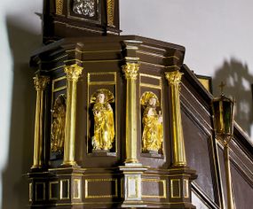 Zdjęcie nr 1: Ambona drewniana, przyścienna, wisząca, dostawiona do północnej ściany nawy ze schodkami biegnącymi wzdłuż ściany. Kosz ambony jest sześcioboczny, z niewielkim parapetem. Jego boki zostały podzielone na pola stojącymi na wysokich cokołach pięcioma kolumienkami o korynckich kapitelach, dźwigających profilowane belkowanie. W polach prostokątne płyciny z konchowymi wnękami ozdobionymi muszlami z figurami ewangelistów. 
We wnękach rzeźby z przedstawieniami ewangelistów i Chrystusa. Figury drewniane pełnoplastyczne, wydrążone od tyłu, polichromowane, złocone i srebrzone, ustawione na niewielkich prostokątnych podstawach, od lewej: św. Łukasz wsparty prawą nogą na głowie byka, św. Marek wsparty prawą nogą na głowie lwa, św. Jan z orłem przy prawej nodze, Chrystus Salvator Mundi, św. Mateusz z aniołem przy prawej nodze. Ewangeliści trzymają w prawych dłoniach otwarte księgi, z wyjątkiem św. Mateusza, trzymającego kodeks w lewej ręce. Dłonie ewangelistów nieproporcjonalnie duże. Szaty bogato pofałdowane, głęboko cięte. 
Podstawa kosza bogato profilowana, ozdobiona podwieszoną dekoracyjną szyszką. Schodki ograniczone parapetem z trzema romboidalnymi płycinami w złoconych profilowanych obramieniach. Na filarze przy wejściu na schodki złocona kula z szyszką. Zaplecek w kształcie stojącego prostokąta, w górnej części ujęty zwężającymi się ku dołowi pilastrami wypełnionymi cekinami, zwieńczonymi rombowymi wypustkami i korynckimi kapitelami, które podtrzymują przełamujące się nad nimi belkowanie. Pośrodku zaplecka w prostokątnym obramieniu zamkniętym nadwieszonym łukiem półkolistym płaskorzeźbione popiersie Boga Ojca wyłaniającego się z obłoków. Prawa ręka Boga wzniesiona w geście błogosławieństwa, lewa oparta na globie ziemskim zwieńczonym krzyżem. Tłem dla postaci Boga Ojca jest rozwiana poła płaszcza układająca się niemal półkoliście. Baldachim ośmioboczny. W profilowanym podniebiu, ozdobionym pasem ornamentu małżowinowo-chrząstkowego i profilowanymi wisiorami, gołębica Ducha Świętego na tle glorii utworzonej z prostych promieni o równej długości. Baldachim ozdobiony zamkniętą koroną, którą tworzy pięć esownic pokrytych pasami ornamentu małżowinowo-chrząstkowego. W zwieńczeniu pełnoplastyczna rzeźba przedstawiająca św. Jana Chrzciciela. Figura drewniana, wydrążona z tyłu, polichromowana, złocona i srebrzona, ustawiona na niewielkiej wielobocznej profilowanej podstawie. Święty w prawej ręce trzyma wysoki krzyż, w lewej księgę, na której spoczywa Baranek Boży. Struktura ambony ciemnobrązowa. Kolumny, kapitele, pilastry w zaplecku korona baldachimu, profilowania i ornamenty złocone, z wyjątkiem srebrzonej ramy w zaplecku i gołębicy Ducha Świętego.