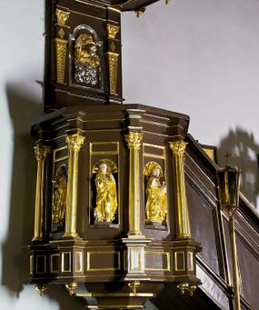 Zdjęcie nr 1: Ambona drewniana, przyścienna, wisząca, dostawiona do północnej ściany nawy ze schodkami biegnącymi wzdłuż ściany. Kosz ambony jest sześcioboczny, z niewielkim parapetem. Jego boki zostały podzielone na pola stojącymi na wysokich cokołach pięcioma kolumienkami o korynckich kapitelach, dźwigających profilowane belkowanie. W polach prostokątne płyciny z konchowymi wnękami ozdobionymi muszlami z figurami ewangelistów. 
We wnękach rzeźby z przedstawieniami ewangelistów i Chrystusa. Figury drewniane pełnoplastyczne, wydrążone od tyłu, polichromowane, złocone i srebrzone, ustawione na niewielkich prostokątnych podstawach, od lewej: św. Łukasz wsparty prawą nogą na głowie byka, św. Marek wsparty prawą nogą na głowie lwa, św. Jan z orłem przy prawej nodze, Chrystus Salvator Mundi, św. Mateusz z aniołem przy prawej nodze. Ewangeliści trzymają w prawych dłoniach otwarte księgi, z wyjątkiem św. Mateusza, trzymającego kodeks w lewej ręce. Dłonie ewangelistów nieproporcjonalnie duże. Szaty bogato pofałdowane, głęboko cięte. 
Podstawa kosza bogato profilowana, ozdobiona podwieszoną dekoracyjną szyszką. Schodki ograniczone parapetem z trzema romboidalnymi płycinami w złoconych profilowanych obramieniach. Na filarze przy wejściu na schodki złocona kula z szyszką. Zaplecek w kształcie stojącego prostokąta, w górnej części ujęty zwężającymi się ku dołowi pilastrami wypełnionymi cekinami, zwieńczonymi rombowymi wypustkami i korynckimi kapitelami, które podtrzymują przełamujące się nad nimi belkowanie. Pośrodku zaplecka w prostokątnym obramieniu zamkniętym nadwieszonym łukiem półkolistym płaskorzeźbione popiersie Boga Ojca wyłaniającego się z obłoków. Prawa ręka Boga wzniesiona w geście błogosławieństwa, lewa oparta na globie ziemskim zwieńczonym krzyżem. Tłem dla postaci Boga Ojca jest rozwiana poła płaszcza układająca się niemal półkoliście. Baldachim ośmioboczny. W profilowanym podniebiu, ozdobionym pasem ornamentu małżowinowo-chrząstkowego i profilowanymi wisiorami, gołębica Ducha Świętego na tle glorii utworzonej z prostych promieni o równej długości. Baldachim ozdobiony zamkniętą koroną, którą tworzy pięć esownic pokrytych pasami ornamentu małżowinowo-chrząstkowego. W zwieńczeniu pełnoplastyczna rzeźba przedstawiająca św. Jana Chrzciciela. Figura drewniana, wydrążona z tyłu, polichromowana, złocona i srebrzona, ustawiona na niewielkiej wielobocznej profilowanej podstawie. Święty w prawej ręce trzyma wysoki krzyż, w lewej księgę, na której spoczywa Baranek Boży. Struktura ambony ciemnobrązowa. Kolumny, kapitele, pilastry w zaplecku korona baldachimu, profilowania i ornamenty złocone, z wyjątkiem srebrzonej ramy w zaplecku i gołębicy Ducha Świętego.