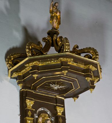 Zdjęcie nr 2: Ambona drewniana, przyścienna, wisząca, dostawiona do północnej ściany nawy ze schodkami biegnącymi wzdłuż ściany. Kosz ambony jest sześcioboczny, z niewielkim parapetem. Jego boki zostały podzielone na pola stojącymi na wysokich cokołach pięcioma kolumienkami o korynckich kapitelach, dźwigających profilowane belkowanie. W polach prostokątne płyciny z konchowymi wnękami ozdobionymi muszlami z figurami ewangelistów. 
We wnękach rzeźby z przedstawieniami ewangelistów i Chrystusa. Figury drewniane pełnoplastyczne, wydrążone od tyłu, polichromowane, złocone i srebrzone, ustawione na niewielkich prostokątnych podstawach, od lewej: św. Łukasz wsparty prawą nogą na głowie byka, św. Marek wsparty prawą nogą na głowie lwa, św. Jan z orłem przy prawej nodze, Chrystus Salvator Mundi, św. Mateusz z aniołem przy prawej nodze. Ewangeliści trzymają w prawych dłoniach otwarte księgi, z wyjątkiem św. Mateusza, trzymającego kodeks w lewej ręce. Dłonie ewangelistów nieproporcjonalnie duże. Szaty bogato pofałdowane, głęboko cięte. 
Podstawa kosza bogato profilowana, ozdobiona podwieszoną dekoracyjną szyszką. Schodki ograniczone parapetem z trzema romboidalnymi płycinami w złoconych profilowanych obramieniach. Na filarze przy wejściu na schodki złocona kula z szyszką. Zaplecek w kształcie stojącego prostokąta, w górnej części ujęty zwężającymi się ku dołowi pilastrami wypełnionymi cekinami, zwieńczonymi rombowymi wypustkami i korynckimi kapitelami, które podtrzymują przełamujące się nad nimi belkowanie. Pośrodku zaplecka w prostokątnym obramieniu zamkniętym nadwieszonym łukiem półkolistym płaskorzeźbione popiersie Boga Ojca wyłaniającego się z obłoków. Prawa ręka Boga wzniesiona w geście błogosławieństwa, lewa oparta na globie ziemskim zwieńczonym krzyżem. Tłem dla postaci Boga Ojca jest rozwiana poła płaszcza układająca się niemal półkoliście. Baldachim ośmioboczny. W profilowanym podniebiu, ozdobionym pasem ornamentu małżowinowo-chrząstkowego i profilowanymi wisiorami, gołębica Ducha Świętego na tle glorii utworzonej z prostych promieni o równej długości. Baldachim ozdobiony zamkniętą koroną, którą tworzy pięć esownic pokrytych pasami ornamentu małżowinowo-chrząstkowego. W zwieńczeniu pełnoplastyczna rzeźba przedstawiająca św. Jana Chrzciciela. Figura drewniana, wydrążona z tyłu, polichromowana, złocona i srebrzona, ustawiona na niewielkiej wielobocznej profilowanej podstawie. Święty w prawej ręce trzyma wysoki krzyż, w lewej księgę, na której spoczywa Baranek Boży. Struktura ambony ciemnobrązowa. Kolumny, kapitele, pilastry w zaplecku korona baldachimu, profilowania i ornamenty złocone, z wyjątkiem srebrzonej ramy w zaplecku i gołębicy Ducha Świętego.