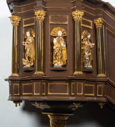 Zdjęcie nr 25: Ambona drewniana, przyścienna, wisząca, dostawiona do północnej ściany nawy ze schodkami biegnącymi wzdłuż ściany. Kosz ambony jest sześcioboczny, z niewielkim parapetem. Jego boki zostały podzielone na pola stojącymi na wysokich cokołach pięcioma kolumienkami o korynckich kapitelach, dźwigających profilowane belkowanie. W polach prostokątne płyciny z konchowymi wnękami ozdobionymi muszlami z figurami ewangelistów. 
We wnękach rzeźby z przedstawieniami ewangelistów i Chrystusa. Figury drewniane pełnoplastyczne, wydrążone od tyłu, polichromowane, złocone i srebrzone, ustawione na niewielkich prostokątnych podstawach, od lewej: św. Łukasz wsparty prawą nogą na głowie byka, św. Marek wsparty prawą nogą na głowie lwa, św. Jan z orłem przy prawej nodze, Chrystus Salvator Mundi, św. Mateusz z aniołem przy prawej nodze. Ewangeliści trzymają w prawych dłoniach otwarte księgi, z wyjątkiem św. Mateusza, trzymającego kodeks w lewej ręce. Dłonie ewangelistów nieproporcjonalnie duże. Szaty bogato pofałdowane, głęboko cięte. 
Podstawa kosza bogato profilowana, ozdobiona podwieszoną dekoracyjną szyszką. Schodki ograniczone parapetem z trzema romboidalnymi płycinami w złoconych profilowanych obramieniach. Na filarze przy wejściu na schodki złocona kula z szyszką. Zaplecek w kształcie stojącego prostokąta, w górnej części ujęty zwężającymi się ku dołowi pilastrami wypełnionymi cekinami, zwieńczonymi rombowymi wypustkami i korynckimi kapitelami, które podtrzymują przełamujące się nad nimi belkowanie. Pośrodku zaplecka w prostokątnym obramieniu zamkniętym nadwieszonym łukiem półkolistym płaskorzeźbione popiersie Boga Ojca wyłaniającego się z obłoków. Prawa ręka Boga wzniesiona w geście błogosławieństwa, lewa oparta na globie ziemskim zwieńczonym krzyżem. Tłem dla postaci Boga Ojca jest rozwiana poła płaszcza układająca się niemal półkoliście. Baldachim ośmioboczny. W profilowanym podniebiu, ozdobionym pasem ornamentu małżowinowo-chrząstkowego i profilowanymi wisiorami, gołębica Ducha Świętego na tle glorii utworzonej z prostych promieni o równej długości. Baldachim ozdobiony zamkniętą koroną, którą tworzy pięć esownic pokrytych pasami ornamentu małżowinowo-chrząstkowego. W zwieńczeniu pełnoplastyczna rzeźba przedstawiająca św. Jana Chrzciciela. Figura drewniana, wydrążona z tyłu, polichromowana, złocona i srebrzona, ustawiona na niewielkiej wielobocznej profilowanej podstawie. Święty w prawej ręce trzyma wysoki krzyż, w lewej księgę, na której spoczywa Baranek Boży. Struktura ambony ciemnobrązowa. Kolumny, kapitele, pilastry w zaplecku korona baldachimu, profilowania i ornamenty złocone, z wyjątkiem srebrzonej ramy w zaplecku i gołębicy Ducha Świętego.