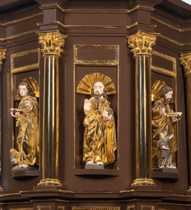 Zdjęcie nr 4: Ambona drewniana, przyścienna, wisząca, dostawiona do północnej ściany nawy ze schodkami biegnącymi wzdłuż ściany. Kosz ambony jest sześcioboczny, z niewielkim parapetem. Jego boki zostały podzielone na pola stojącymi na wysokich cokołach pięcioma kolumienkami o korynckich kapitelach, dźwigających profilowane belkowanie. W polach prostokątne płyciny z konchowymi wnękami ozdobionymi muszlami z figurami ewangelistów. 
We wnękach rzeźby z przedstawieniami ewangelistów i Chrystusa. Figury drewniane pełnoplastyczne, wydrążone od tyłu, polichromowane, złocone i srebrzone, ustawione na niewielkich prostokątnych podstawach, od lewej: św. Łukasz wsparty prawą nogą na głowie byka, św. Marek wsparty prawą nogą na głowie lwa, św. Jan z orłem przy prawej nodze, Chrystus Salvator Mundi, św. Mateusz z aniołem przy prawej nodze. Ewangeliści trzymają w prawych dłoniach otwarte księgi, z wyjątkiem św. Mateusza, trzymającego kodeks w lewej ręce. Dłonie ewangelistów nieproporcjonalnie duże. Szaty bogato pofałdowane, głęboko cięte. 
Podstawa kosza bogato profilowana, ozdobiona podwieszoną dekoracyjną szyszką. Schodki ograniczone parapetem z trzema romboidalnymi płycinami w złoconych profilowanych obramieniach. Na filarze przy wejściu na schodki złocona kula z szyszką. Zaplecek w kształcie stojącego prostokąta, w górnej części ujęty zwężającymi się ku dołowi pilastrami wypełnionymi cekinami, zwieńczonymi rombowymi wypustkami i korynckimi kapitelami, które podtrzymują przełamujące się nad nimi belkowanie. Pośrodku zaplecka w prostokątnym obramieniu zamkniętym nadwieszonym łukiem półkolistym płaskorzeźbione popiersie Boga Ojca wyłaniającego się z obłoków. Prawa ręka Boga wzniesiona w geście błogosławieństwa, lewa oparta na globie ziemskim zwieńczonym krzyżem. Tłem dla postaci Boga Ojca jest rozwiana poła płaszcza układająca się niemal półkoliście. Baldachim ośmioboczny. W profilowanym podniebiu, ozdobionym pasem ornamentu małżowinowo-chrząstkowego i profilowanymi wisiorami, gołębica Ducha Świętego na tle glorii utworzonej z prostych promieni o równej długości. Baldachim ozdobiony zamkniętą koroną, którą tworzy pięć esownic pokrytych pasami ornamentu małżowinowo-chrząstkowego. W zwieńczeniu pełnoplastyczna rzeźba przedstawiająca św. Jana Chrzciciela. Figura drewniana, wydrążona z tyłu, polichromowana, złocona i srebrzona, ustawiona na niewielkiej wielobocznej profilowanej podstawie. Święty w prawej ręce trzyma wysoki krzyż, w lewej księgę, na której spoczywa Baranek Boży. Struktura ambony ciemnobrązowa. Kolumny, kapitele, pilastry w zaplecku korona baldachimu, profilowania i ornamenty złocone, z wyjątkiem srebrzonej ramy w zaplecku i gołębicy Ducha Świętego.