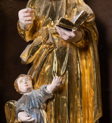 Zdjęcie nr 5: Ambona drewniana, przyścienna, wisząca, dostawiona do północnej ściany nawy ze schodkami biegnącymi wzdłuż ściany. Kosz ambony jest sześcioboczny, z niewielkim parapetem. Jego boki zostały podzielone na pola stojącymi na wysokich cokołach pięcioma kolumienkami o korynckich kapitelach, dźwigających profilowane belkowanie. W polach prostokątne płyciny z konchowymi wnękami ozdobionymi muszlami z figurami ewangelistów. 
We wnękach rzeźby z przedstawieniami ewangelistów i Chrystusa. Figury drewniane pełnoplastyczne, wydrążone od tyłu, polichromowane, złocone i srebrzone, ustawione na niewielkich prostokątnych podstawach, od lewej: św. Łukasz wsparty prawą nogą na głowie byka, św. Marek wsparty prawą nogą na głowie lwa, św. Jan z orłem przy prawej nodze, Chrystus Salvator Mundi, św. Mateusz z aniołem przy prawej nodze. Ewangeliści trzymają w prawych dłoniach otwarte księgi, z wyjątkiem św. Mateusza, trzymającego kodeks w lewej ręce. Dłonie ewangelistów nieproporcjonalnie duże. Szaty bogato pofałdowane, głęboko cięte. 
Podstawa kosza bogato profilowana, ozdobiona podwieszoną dekoracyjną szyszką. Schodki ograniczone parapetem z trzema romboidalnymi płycinami w złoconych profilowanych obramieniach. Na filarze przy wejściu na schodki złocona kula z szyszką. Zaplecek w kształcie stojącego prostokąta, w górnej części ujęty zwężającymi się ku dołowi pilastrami wypełnionymi cekinami, zwieńczonymi rombowymi wypustkami i korynckimi kapitelami, które podtrzymują przełamujące się nad nimi belkowanie. Pośrodku zaplecka w prostokątnym obramieniu zamkniętym nadwieszonym łukiem półkolistym płaskorzeźbione popiersie Boga Ojca wyłaniającego się z obłoków. Prawa ręka Boga wzniesiona w geście błogosławieństwa, lewa oparta na globie ziemskim zwieńczonym krzyżem. Tłem dla postaci Boga Ojca jest rozwiana poła płaszcza układająca się niemal półkoliście. Baldachim ośmioboczny. W profilowanym podniebiu, ozdobionym pasem ornamentu małżowinowo-chrząstkowego i profilowanymi wisiorami, gołębica Ducha Świętego na tle glorii utworzonej z prostych promieni o równej długości. Baldachim ozdobiony zamkniętą koroną, którą tworzy pięć esownic pokrytych pasami ornamentu małżowinowo-chrząstkowego. W zwieńczeniu pełnoplastyczna rzeźba przedstawiająca św. Jana Chrzciciela. Figura drewniana, wydrążona z tyłu, polichromowana, złocona i srebrzona, ustawiona na niewielkiej wielobocznej profilowanej podstawie. Święty w prawej ręce trzyma wysoki krzyż, w lewej księgę, na której spoczywa Baranek Boży. Struktura ambony ciemnobrązowa. Kolumny, kapitele, pilastry w zaplecku korona baldachimu, profilowania i ornamenty złocone, z wyjątkiem srebrzonej ramy w zaplecku i gołębicy Ducha Świętego.