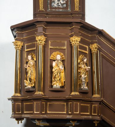 Zdjęcie nr 8: Ambona drewniana, przyścienna, wisząca, dostawiona do północnej ściany nawy ze schodkami biegnącymi wzdłuż ściany. Kosz ambony jest sześcioboczny, z niewielkim parapetem. Jego boki zostały podzielone na pola stojącymi na wysokich cokołach pięcioma kolumienkami o korynckich kapitelach, dźwigających profilowane belkowanie. W polach prostokątne płyciny z konchowymi wnękami ozdobionymi muszlami z figurami ewangelistów. 
We wnękach rzeźby z przedstawieniami ewangelistów i Chrystusa. Figury drewniane pełnoplastyczne, wydrążone od tyłu, polichromowane, złocone i srebrzone, ustawione na niewielkich prostokątnych podstawach, od lewej: św. Łukasz wsparty prawą nogą na głowie byka, św. Marek wsparty prawą nogą na głowie lwa, św. Jan z orłem przy prawej nodze, Chrystus Salvator Mundi, św. Mateusz z aniołem przy prawej nodze. Ewangeliści trzymają w prawych dłoniach otwarte księgi, z wyjątkiem św. Mateusza, trzymającego kodeks w lewej ręce. Dłonie ewangelistów nieproporcjonalnie duże. Szaty bogato pofałdowane, głęboko cięte. 
Podstawa kosza bogato profilowana, ozdobiona podwieszoną dekoracyjną szyszką. Schodki ograniczone parapetem z trzema romboidalnymi płycinami w złoconych profilowanych obramieniach. Na filarze przy wejściu na schodki złocona kula z szyszką. Zaplecek w kształcie stojącego prostokąta, w górnej części ujęty zwężającymi się ku dołowi pilastrami wypełnionymi cekinami, zwieńczonymi rombowymi wypustkami i korynckimi kapitelami, które podtrzymują przełamujące się nad nimi belkowanie. Pośrodku zaplecka w prostokątnym obramieniu zamkniętym nadwieszonym łukiem półkolistym płaskorzeźbione popiersie Boga Ojca wyłaniającego się z obłoków. Prawa ręka Boga wzniesiona w geście błogosławieństwa, lewa oparta na globie ziemskim zwieńczonym krzyżem. Tłem dla postaci Boga Ojca jest rozwiana poła płaszcza układająca się niemal półkoliście. Baldachim ośmioboczny. W profilowanym podniebiu, ozdobionym pasem ornamentu małżowinowo-chrząstkowego i profilowanymi wisiorami, gołębica Ducha Świętego na tle glorii utworzonej z prostych promieni o równej długości. Baldachim ozdobiony zamkniętą koroną, którą tworzy pięć esownic pokrytych pasami ornamentu małżowinowo-chrząstkowego. W zwieńczeniu pełnoplastyczna rzeźba przedstawiająca św. Jana Chrzciciela. Figura drewniana, wydrążona z tyłu, polichromowana, złocona i srebrzona, ustawiona na niewielkiej wielobocznej profilowanej podstawie. Święty w prawej ręce trzyma wysoki krzyż, w lewej księgę, na której spoczywa Baranek Boży. Struktura ambony ciemnobrązowa. Kolumny, kapitele, pilastry w zaplecku korona baldachimu, profilowania i ornamenty złocone, z wyjątkiem srebrzonej ramy w zaplecku i gołębicy Ducha Świętego.