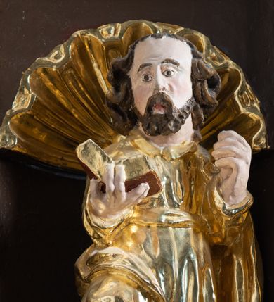 Zdjęcie nr 17: Ambona drewniana, przyścienna, wisząca, dostawiona do północnej ściany nawy ze schodkami biegnącymi wzdłuż ściany. Kosz ambony jest sześcioboczny, z niewielkim parapetem. Jego boki zostały podzielone na pola stojącymi na wysokich cokołach pięcioma kolumienkami o korynckich kapitelach, dźwigających profilowane belkowanie. W polach prostokątne płyciny z konchowymi wnękami ozdobionymi muszlami z figurami ewangelistów. 
We wnękach rzeźby z przedstawieniami ewangelistów i Chrystusa. Figury drewniane pełnoplastyczne, wydrążone od tyłu, polichromowane, złocone i srebrzone, ustawione na niewielkich prostokątnych podstawach, od lewej: św. Łukasz wsparty prawą nogą na głowie byka, św. Marek wsparty prawą nogą na głowie lwa, św. Jan z orłem przy prawej nodze, Chrystus Salvator Mundi, św. Mateusz z aniołem przy prawej nodze. Ewangeliści trzymają w prawych dłoniach otwarte księgi, z wyjątkiem św. Mateusza, trzymającego kodeks w lewej ręce. Dłonie ewangelistów nieproporcjonalnie duże. Szaty bogato pofałdowane, głęboko cięte. 
Podstawa kosza bogato profilowana, ozdobiona podwieszoną dekoracyjną szyszką. Schodki ograniczone parapetem z trzema romboidalnymi płycinami w złoconych profilowanych obramieniach. Na filarze przy wejściu na schodki złocona kula z szyszką. Zaplecek w kształcie stojącego prostokąta, w górnej części ujęty zwężającymi się ku dołowi pilastrami wypełnionymi cekinami, zwieńczonymi rombowymi wypustkami i korynckimi kapitelami, które podtrzymują przełamujące się nad nimi belkowanie. Pośrodku zaplecka w prostokątnym obramieniu zamkniętym nadwieszonym łukiem półkolistym płaskorzeźbione popiersie Boga Ojca wyłaniającego się z obłoków. Prawa ręka Boga wzniesiona w geście błogosławieństwa, lewa oparta na globie ziemskim zwieńczonym krzyżem. Tłem dla postaci Boga Ojca jest rozwiana poła płaszcza układająca się niemal półkoliście. Baldachim ośmioboczny. W profilowanym podniebiu, ozdobionym pasem ornamentu małżowinowo-chrząstkowego i profilowanymi wisiorami, gołębica Ducha Świętego na tle glorii utworzonej z prostych promieni o równej długości. Baldachim ozdobiony zamkniętą koroną, którą tworzy pięć esownic pokrytych pasami ornamentu małżowinowo-chrząstkowego. W zwieńczeniu pełnoplastyczna rzeźba przedstawiająca św. Jana Chrzciciela. Figura drewniana, wydrążona z tyłu, polichromowana, złocona i srebrzona, ustawiona na niewielkiej wielobocznej profilowanej podstawie. Święty w prawej ręce trzyma wysoki krzyż, w lewej księgę, na której spoczywa Baranek Boży. Struktura ambony ciemnobrązowa. Kolumny, kapitele, pilastry w zaplecku korona baldachimu, profilowania i ornamenty złocone, z wyjątkiem srebrzonej ramy w zaplecku i gołębicy Ducha Świętego.