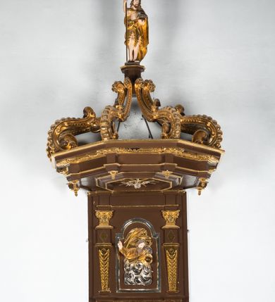 Zdjęcie nr 16: Ambona drewniana, przyścienna, wisząca, dostawiona do północnej ściany nawy ze schodkami biegnącymi wzdłuż ściany. Kosz ambony jest sześcioboczny, z niewielkim parapetem. Jego boki zostały podzielone na pola stojącymi na wysokich cokołach pięcioma kolumienkami o korynckich kapitelach, dźwigających profilowane belkowanie. W polach prostokątne płyciny z konchowymi wnękami ozdobionymi muszlami z figurami ewangelistów. 
We wnękach rzeźby z przedstawieniami ewangelistów i Chrystusa. Figury drewniane pełnoplastyczne, wydrążone od tyłu, polichromowane, złocone i srebrzone, ustawione na niewielkich prostokątnych podstawach, od lewej: św. Łukasz wsparty prawą nogą na głowie byka, św. Marek wsparty prawą nogą na głowie lwa, św. Jan z orłem przy prawej nodze, Chrystus Salvator Mundi, św. Mateusz z aniołem przy prawej nodze. Ewangeliści trzymają w prawych dłoniach otwarte księgi, z wyjątkiem św. Mateusza, trzymającego kodeks w lewej ręce. Dłonie ewangelistów nieproporcjonalnie duże. Szaty bogato pofałdowane, głęboko cięte. 
Podstawa kosza bogato profilowana, ozdobiona podwieszoną dekoracyjną szyszką. Schodki ograniczone parapetem z trzema romboidalnymi płycinami w złoconych profilowanych obramieniach. Na filarze przy wejściu na schodki złocona kula z szyszką. Zaplecek w kształcie stojącego prostokąta, w górnej części ujęty zwężającymi się ku dołowi pilastrami wypełnionymi cekinami, zwieńczonymi rombowymi wypustkami i korynckimi kapitelami, które podtrzymują przełamujące się nad nimi belkowanie. Pośrodku zaplecka w prostokątnym obramieniu zamkniętym nadwieszonym łukiem półkolistym płaskorzeźbione popiersie Boga Ojca wyłaniającego się z obłoków. Prawa ręka Boga wzniesiona w geście błogosławieństwa, lewa oparta na globie ziemskim zwieńczonym krzyżem. Tłem dla postaci Boga Ojca jest rozwiana poła płaszcza układająca się niemal półkoliście. Baldachim ośmioboczny. W profilowanym podniebiu, ozdobionym pasem ornamentu małżowinowo-chrząstkowego i profilowanymi wisiorami, gołębica Ducha Świętego na tle glorii utworzonej z prostych promieni o równej długości. Baldachim ozdobiony zamkniętą koroną, którą tworzy pięć esownic pokrytych pasami ornamentu małżowinowo-chrząstkowego. W zwieńczeniu pełnoplastyczna rzeźba przedstawiająca św. Jana Chrzciciela. Figura drewniana, wydrążona z tyłu, polichromowana, złocona i srebrzona, ustawiona na niewielkiej wielobocznej profilowanej podstawie. Święty w prawej ręce trzyma wysoki krzyż, w lewej księgę, na której spoczywa Baranek Boży. Struktura ambony ciemnobrązowa. Kolumny, kapitele, pilastry w zaplecku korona baldachimu, profilowania i ornamenty złocone, z wyjątkiem srebrzonej ramy w zaplecku i gołębicy Ducha Świętego.